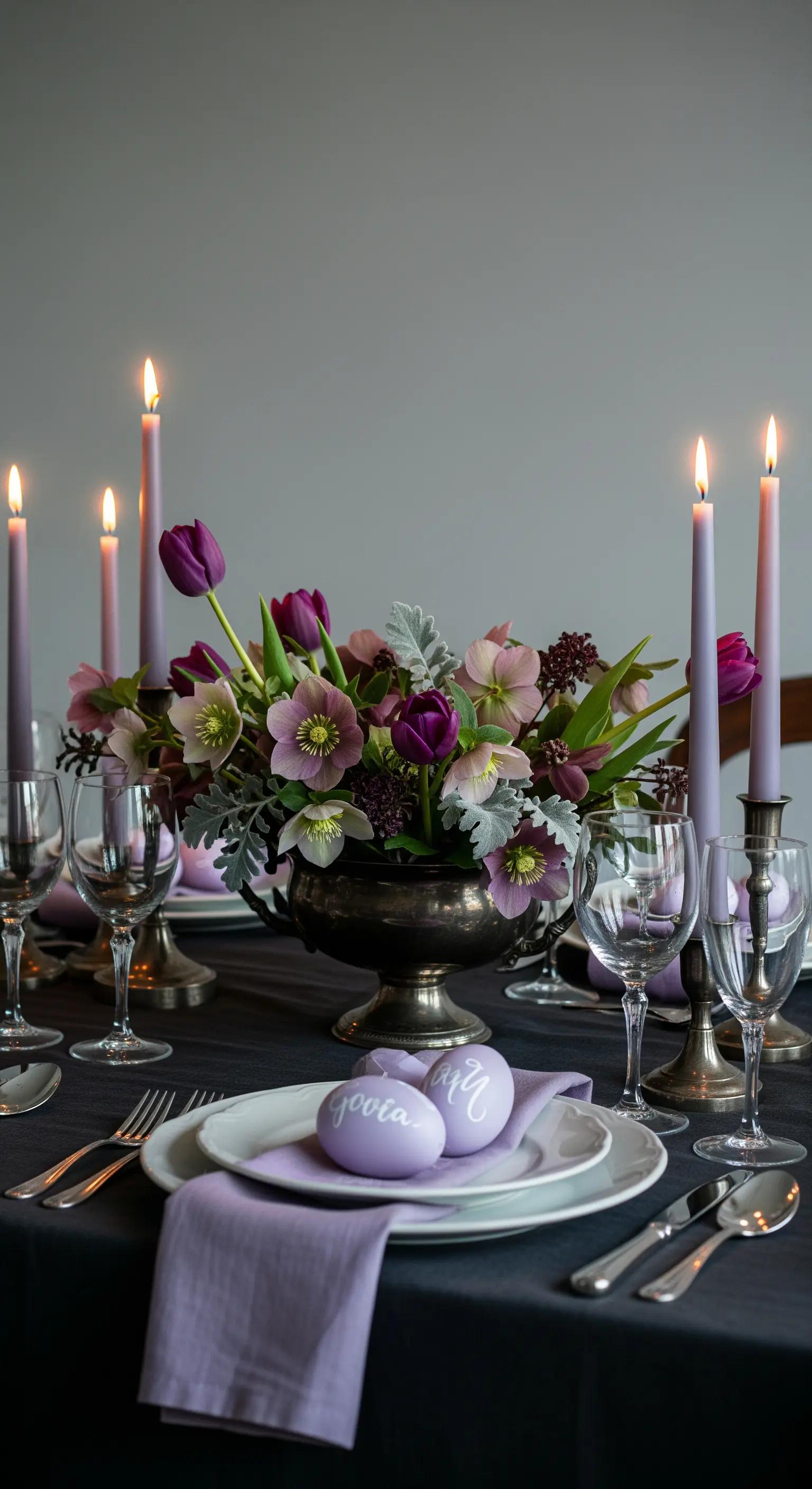 Tavola pasquale scura con fiori viola, candele color lavanda e uova segnaposto.