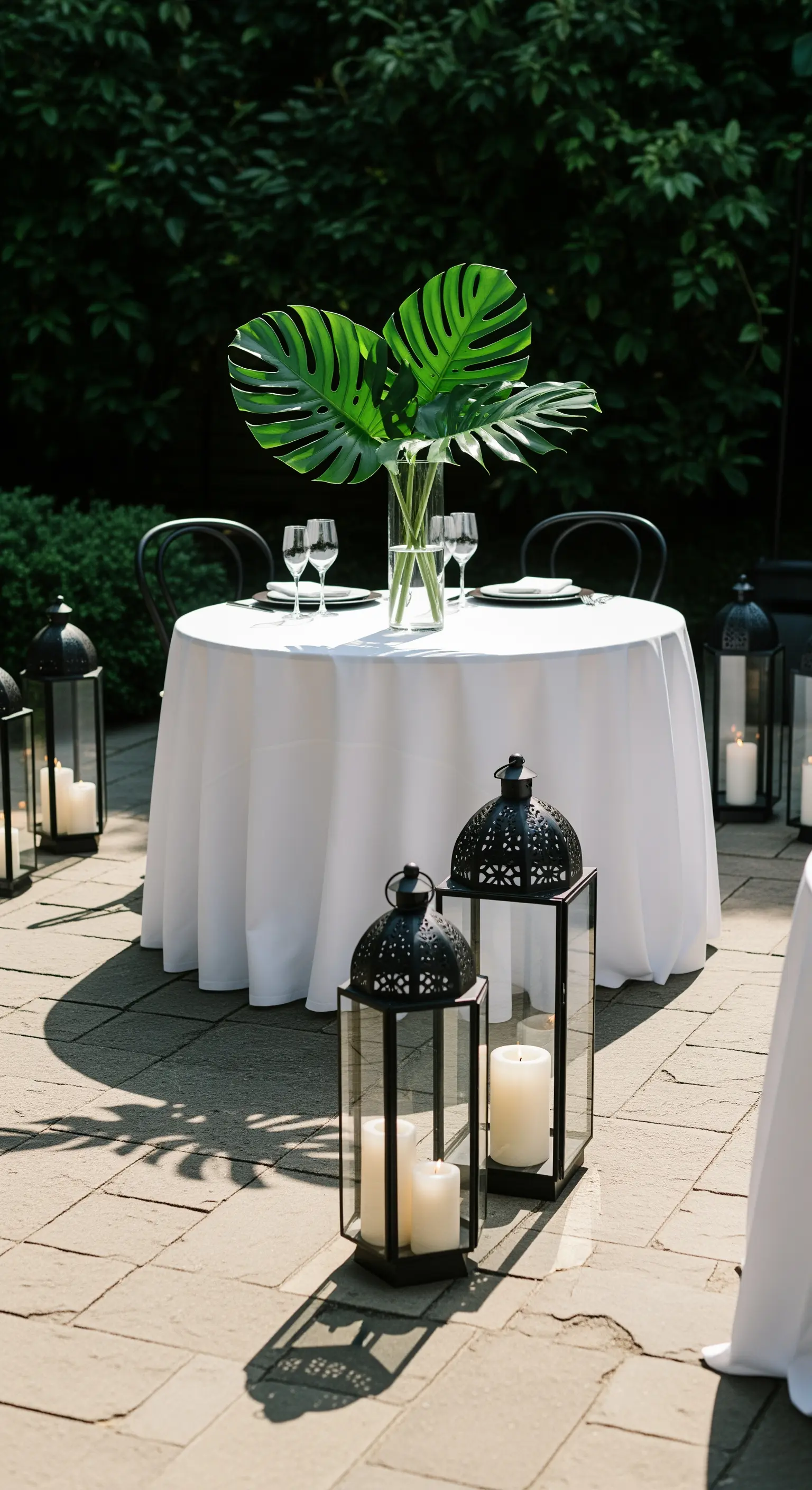 Tavolo rotondo con tovaglia bianca, foglie di Monstera e lanterne nere in un patio.