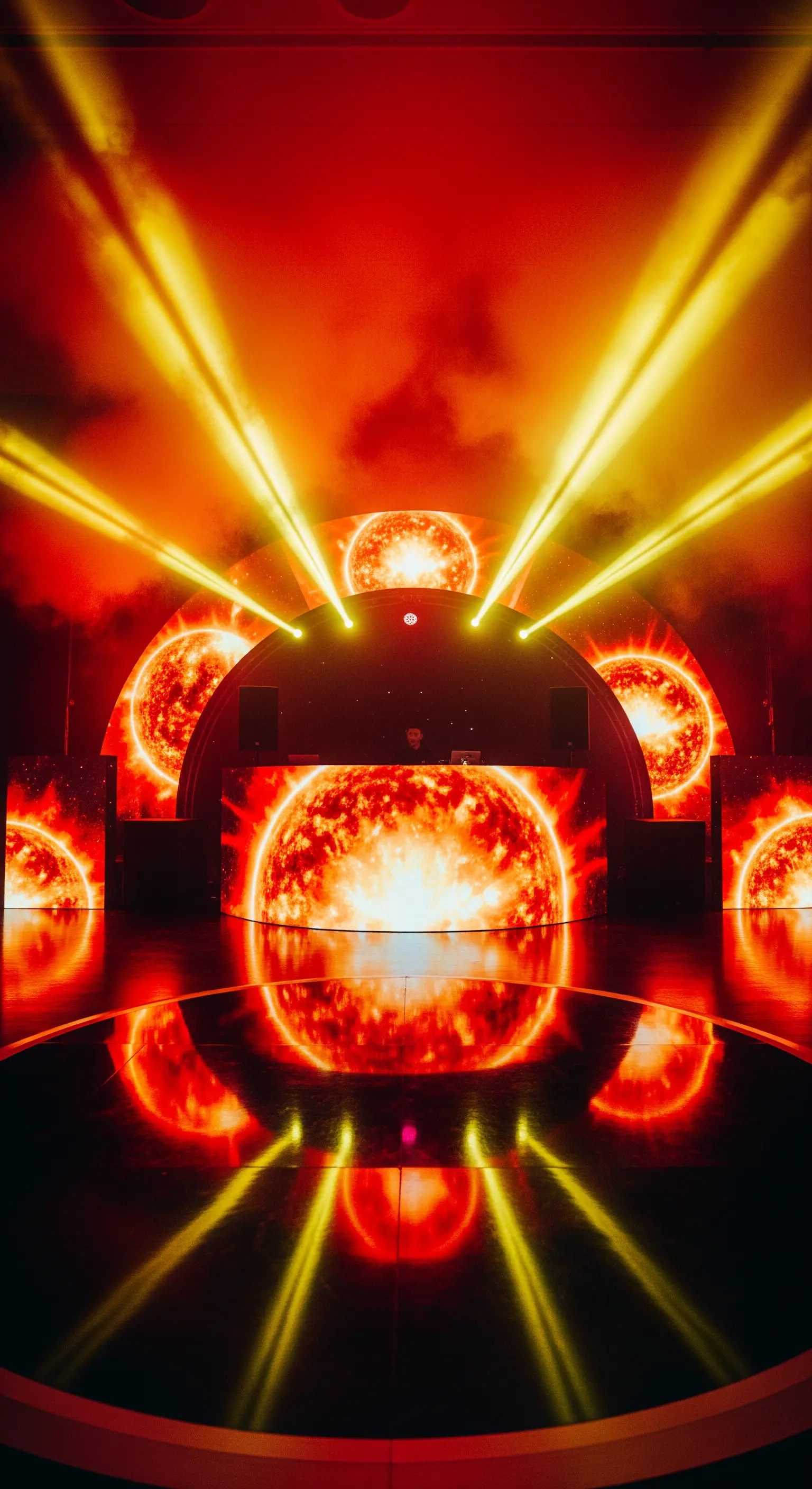 DJ booth circondato da schermi LED che mostrano il sole e fasci di luce gialla.