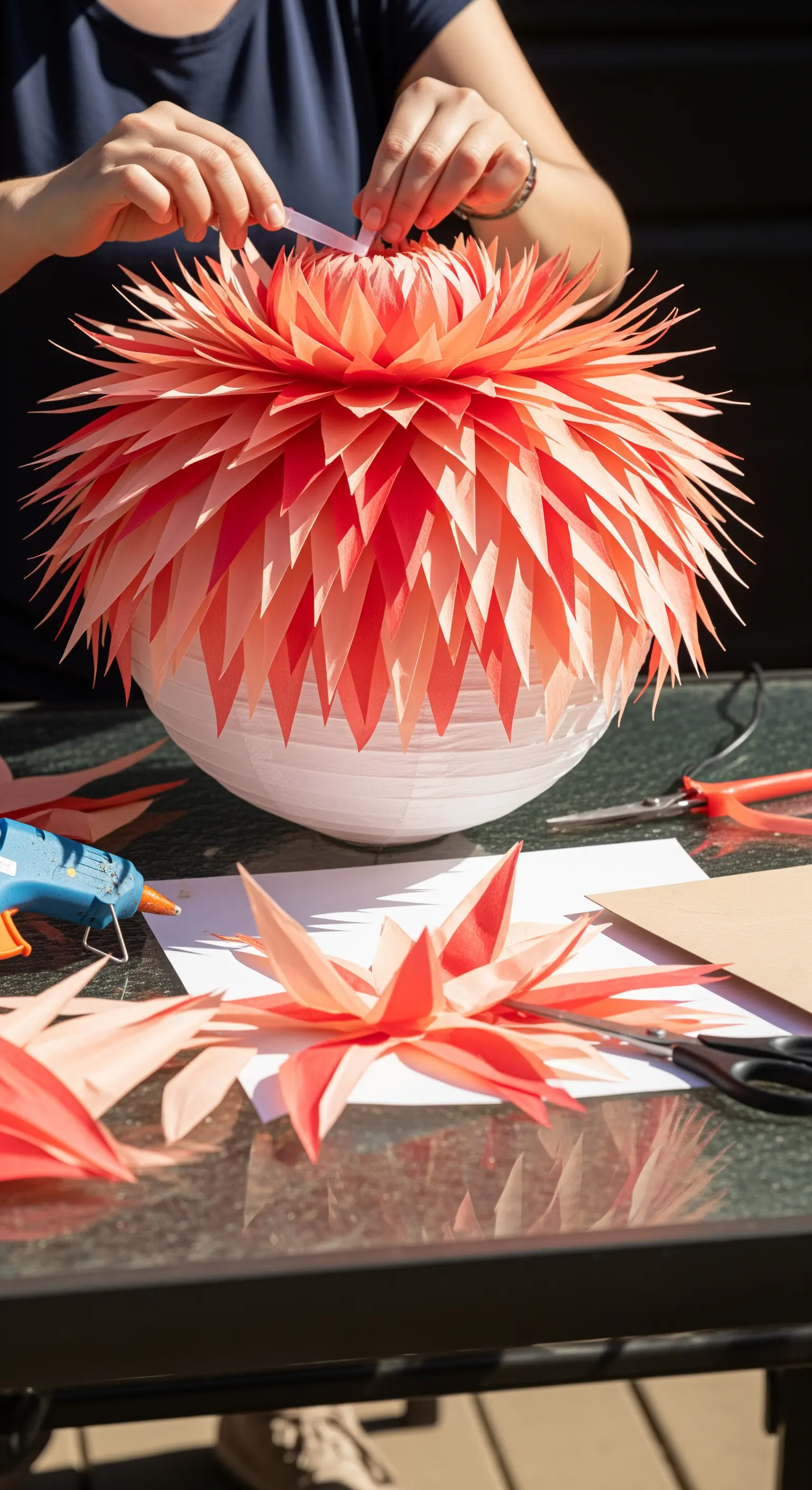 Mani che incollano petali di carta color corallo su una lanterna bianca per creare un fiore 3D.