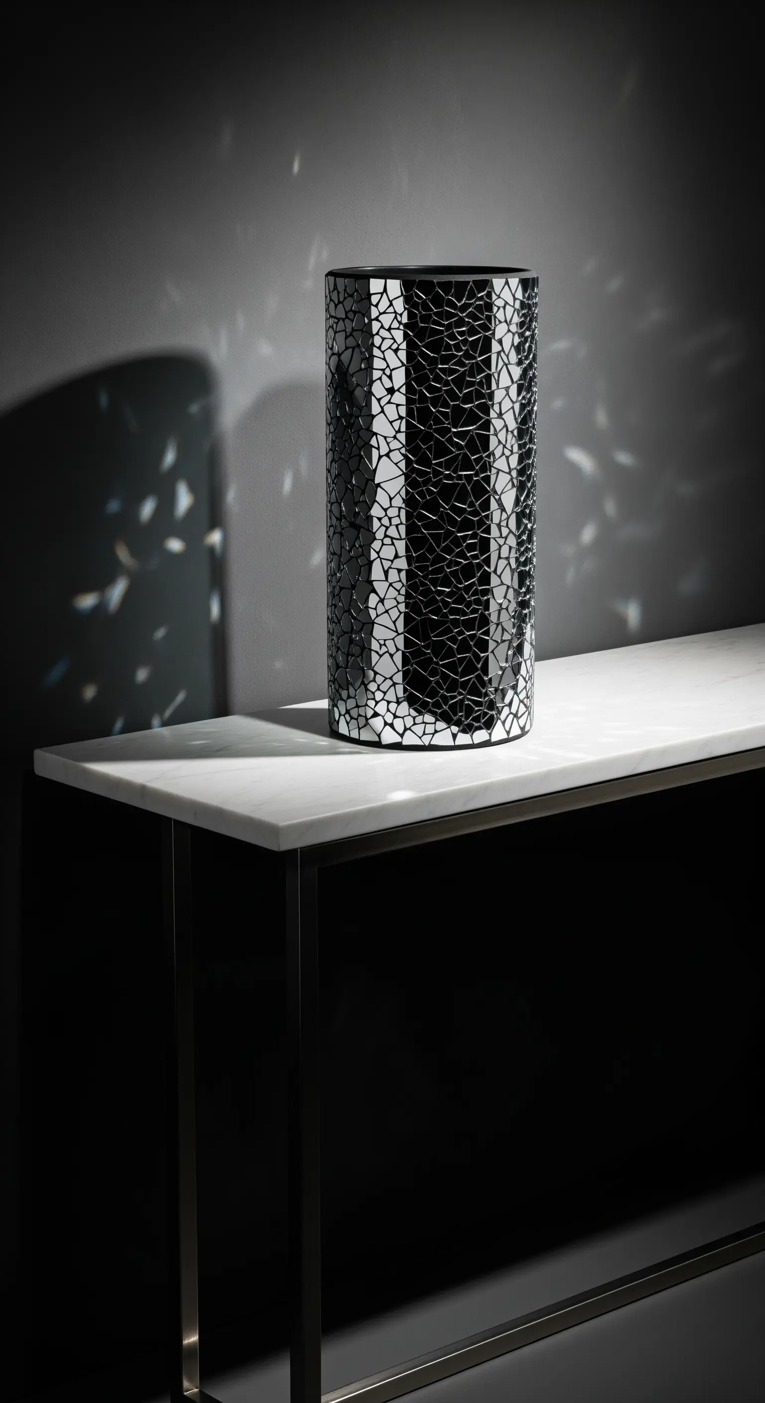 Vaso nero cilindrico con mosaico di specchi rotti, posato su una consolle di marmo in un ambiente buio.