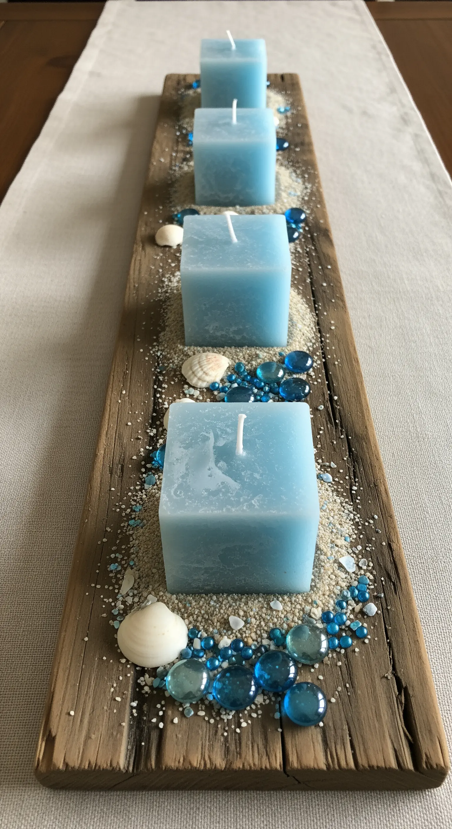 Asse di legno di recupero con candele quadrate azzurre, sabbia e gocce di vetro blu.