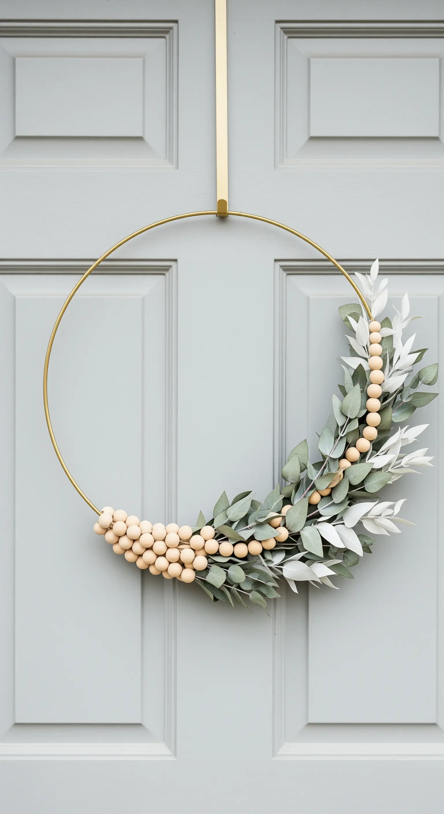 Ghirlanda moderna su cerchio dorato con perline di legno ed eucalipto, appesa a una porta grigia.