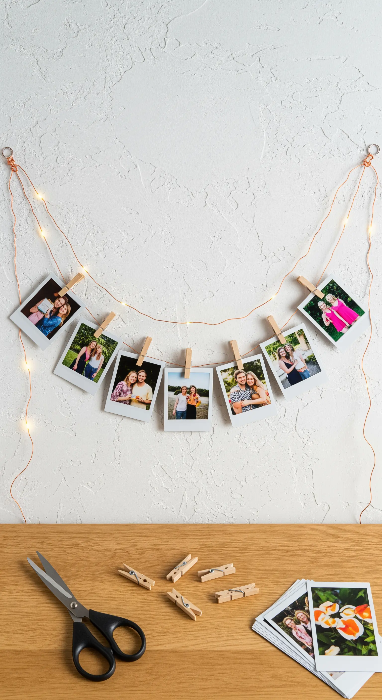 Filo di luci a LED con fotografie stile polaroid appese con mollette di legno su una parete bianca.
