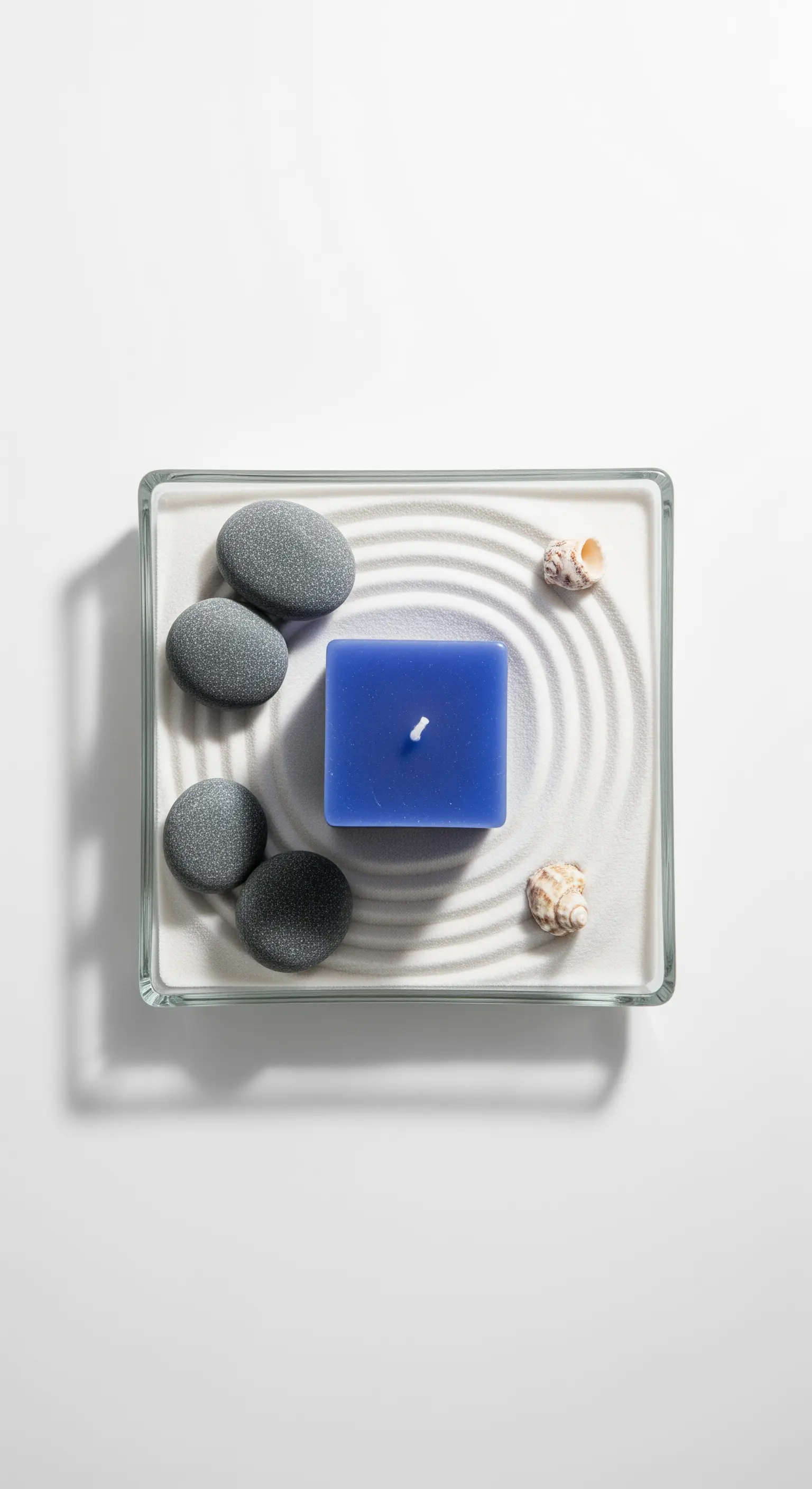 Composizione zen minimalista con candela quadrata blu, sabbia bianca e pietre levigate.