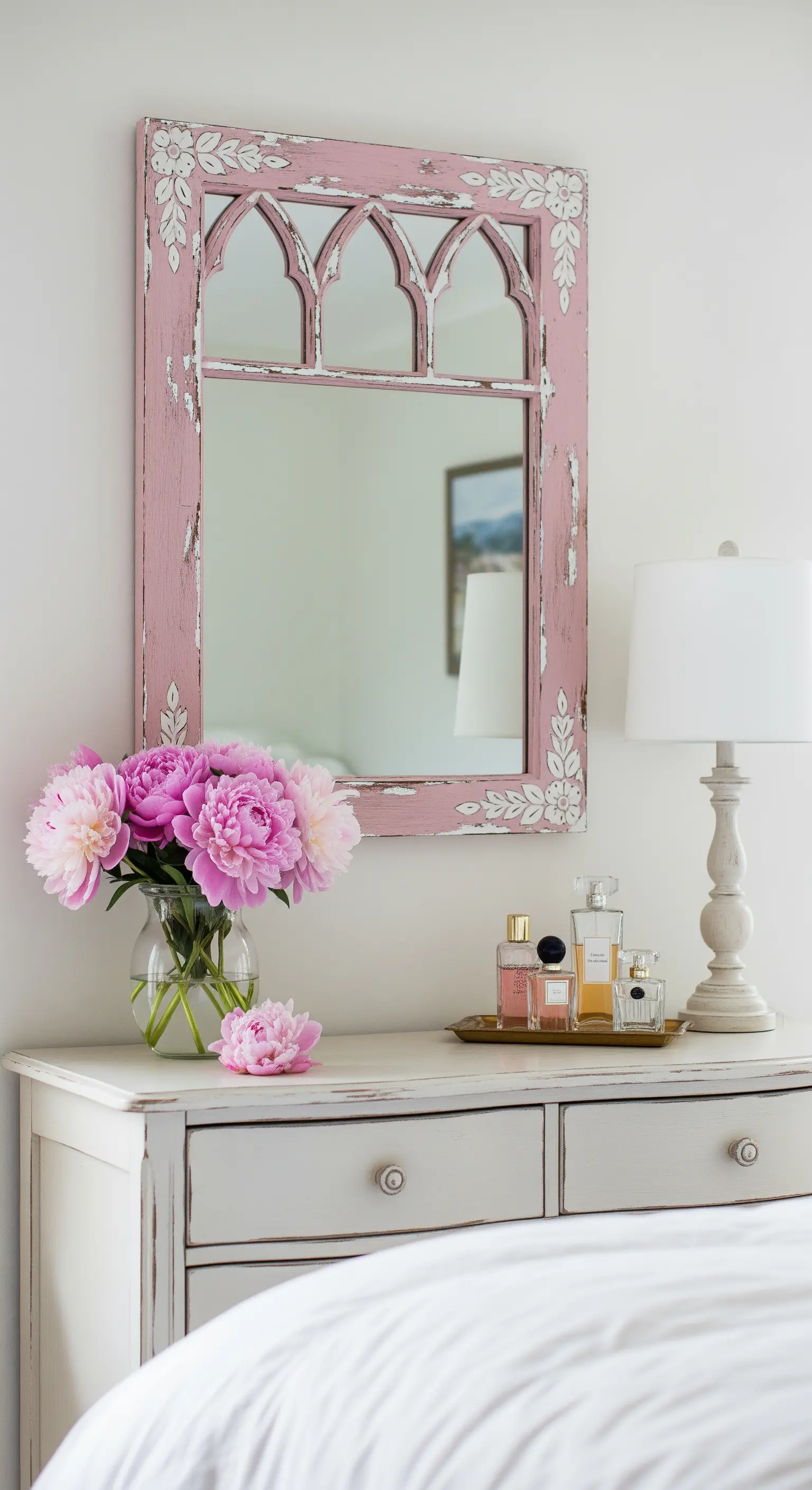 Specchio con cornice rosa a forma di finestra gotica, poggiato su un comò bianco.