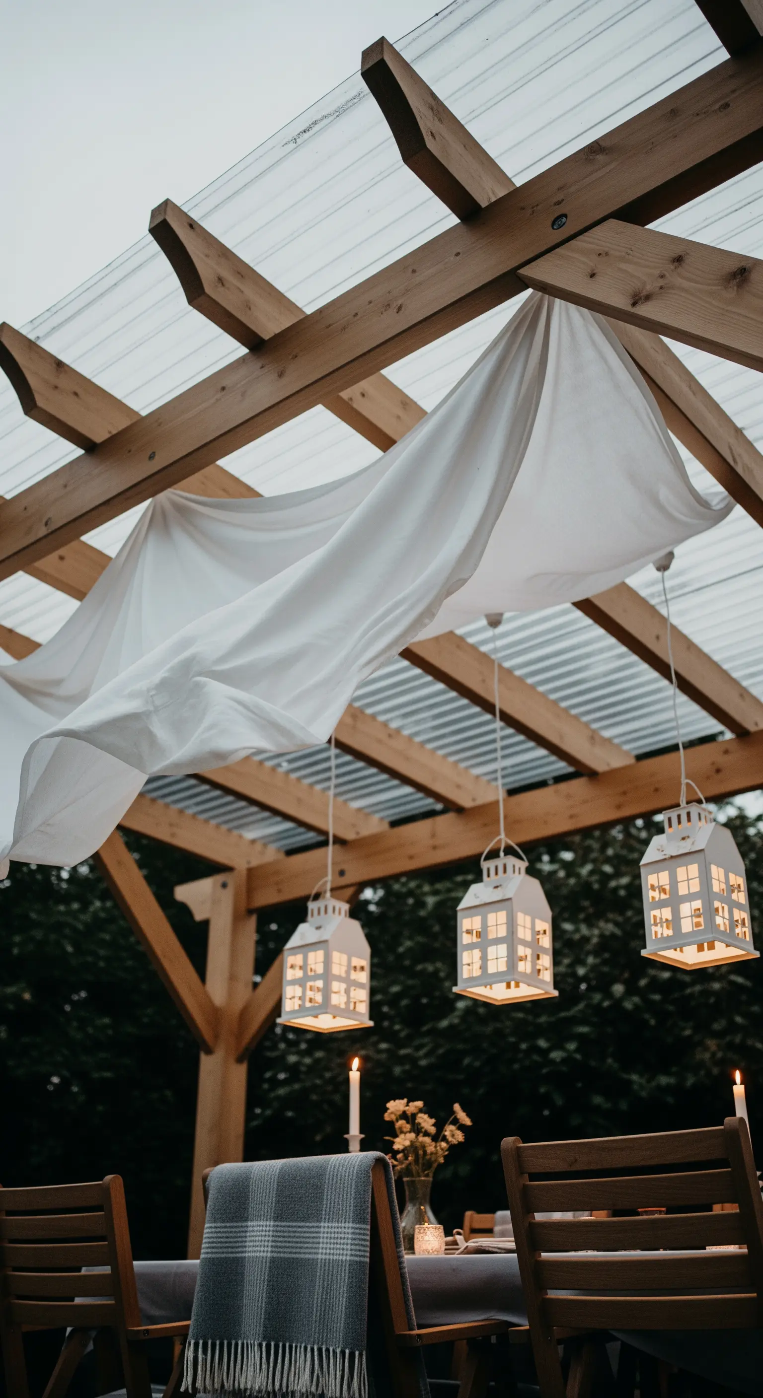 Pergola in legno con tessuto bianco drappeggiato e tre lanterne a forma di casetta sospese.