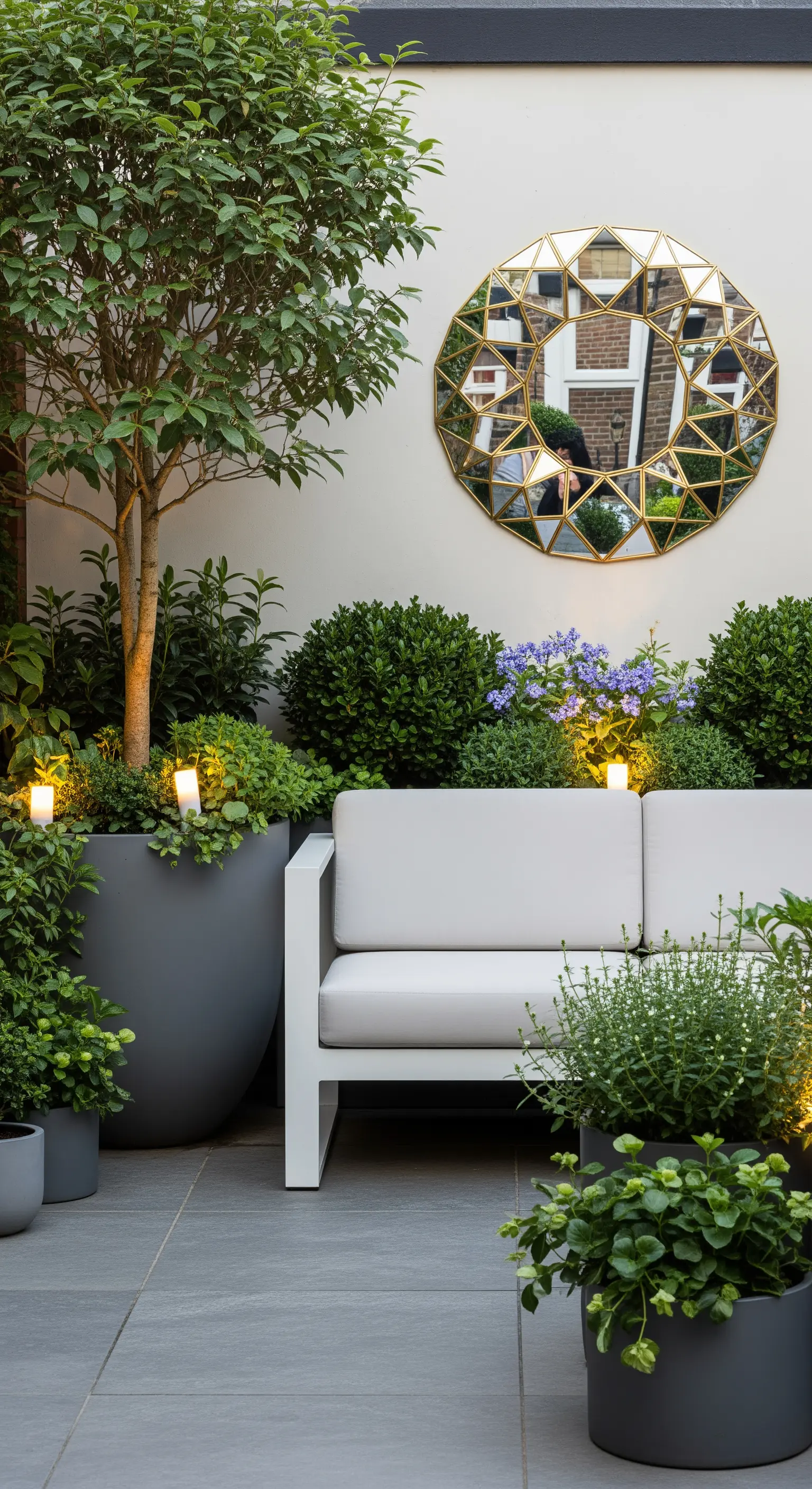 Divano bianco in un piccolo giardino con fioriere, piante e uno specchio geometrico dorato