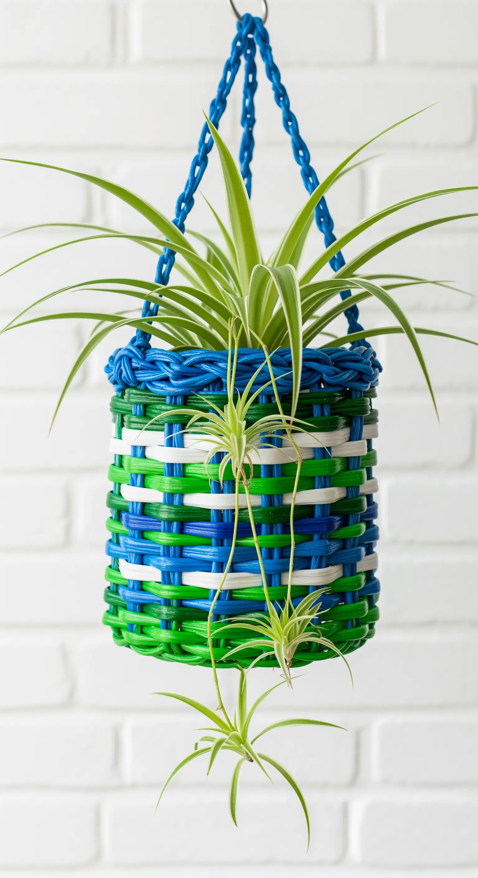 Cesto fioriera sospeso, realizzato intrecciando strisce di plastica blu, verdi e bianche.