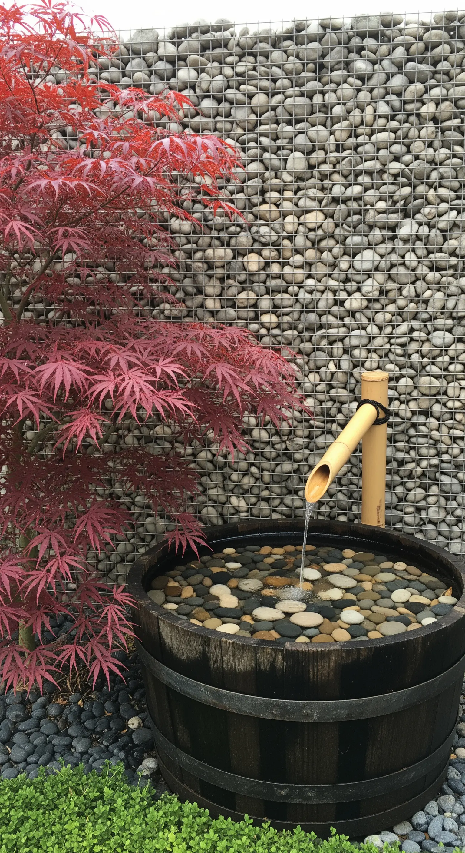 Botte trasformata in una piccola fontana con getto d'acqua in bambù e sassi, accanto a un acero rosso.