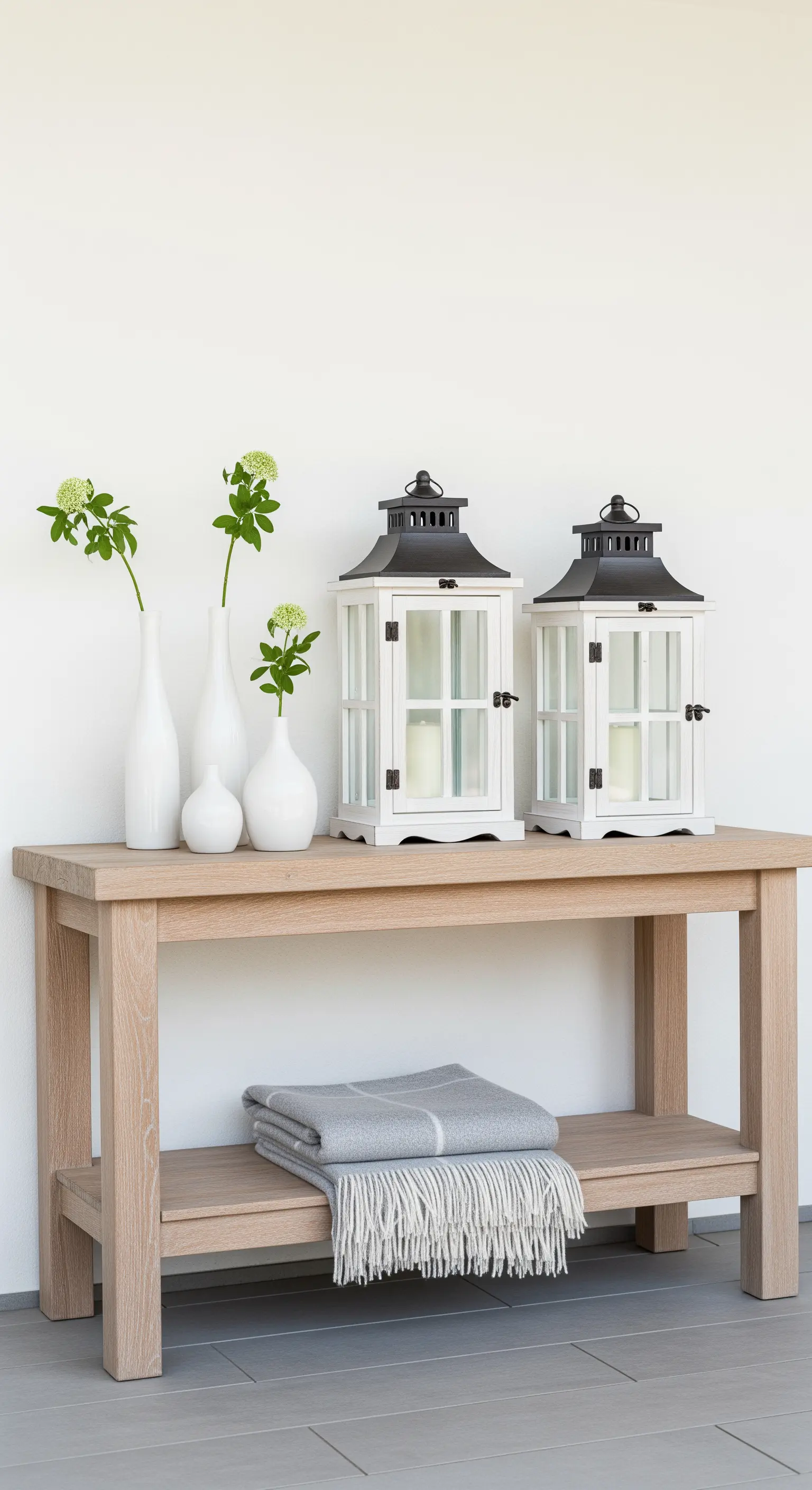 Console in legno chiaro con due lanterne, vasi bianchi e un plaid grigio ripiegato.