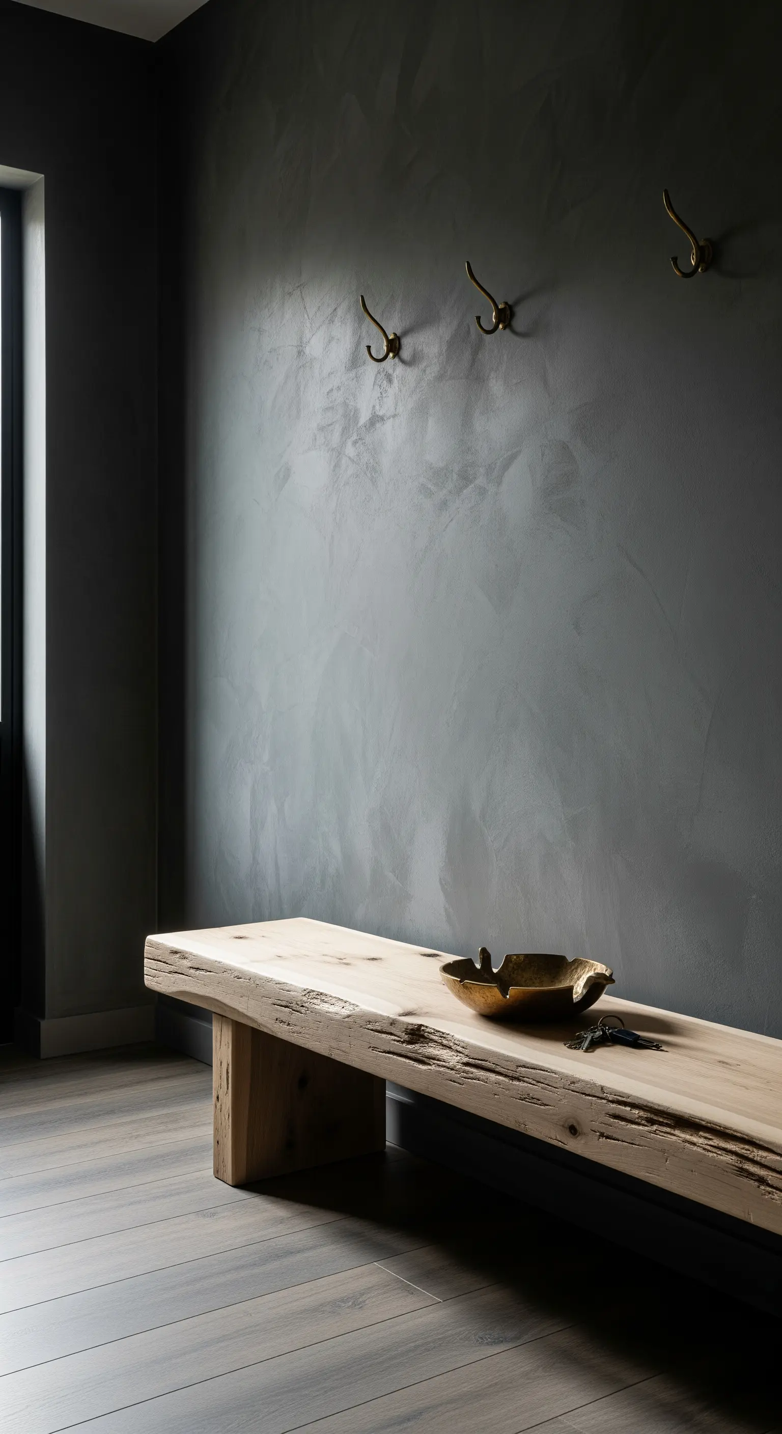Parete materica grigio scuro con panca in legno grezzo e ganci in ottone anticato.