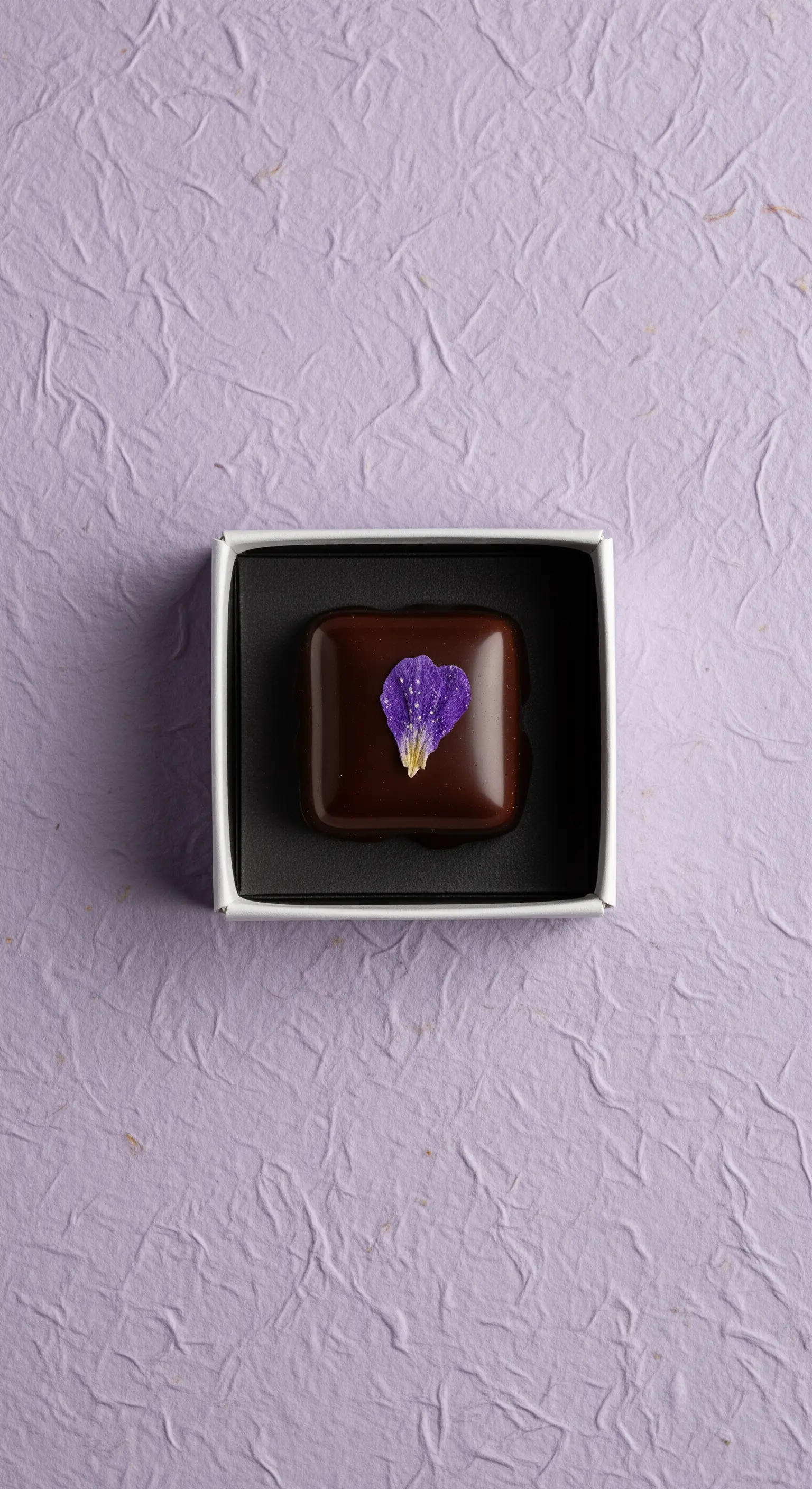 Singolo cioccolatino quadrato decorato con un petalo viola, in una piccola scatola bianca.