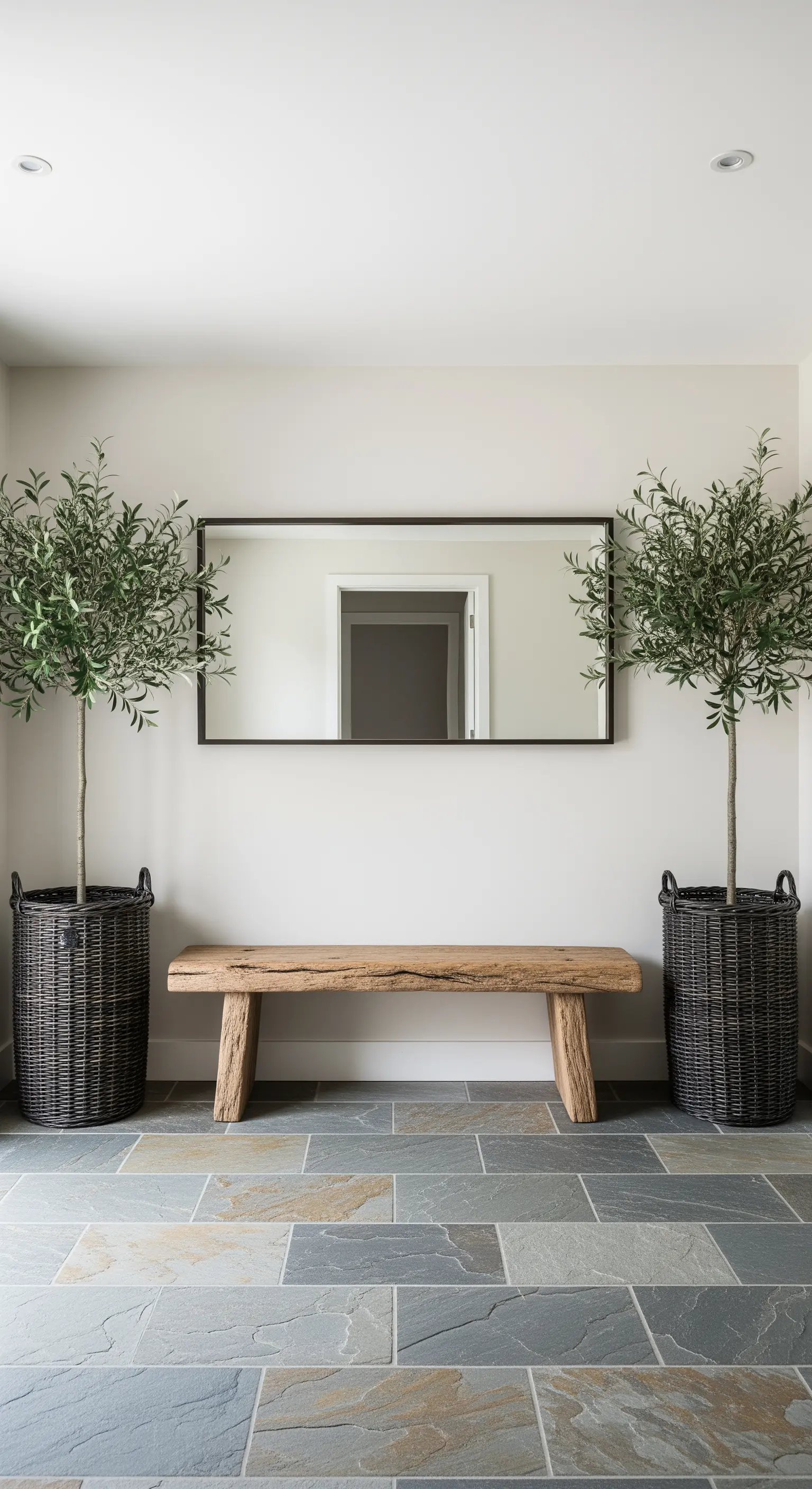 Ingresso simmetrico con panca in legno, specchio orizzontale e due alberi in cesti scuri.