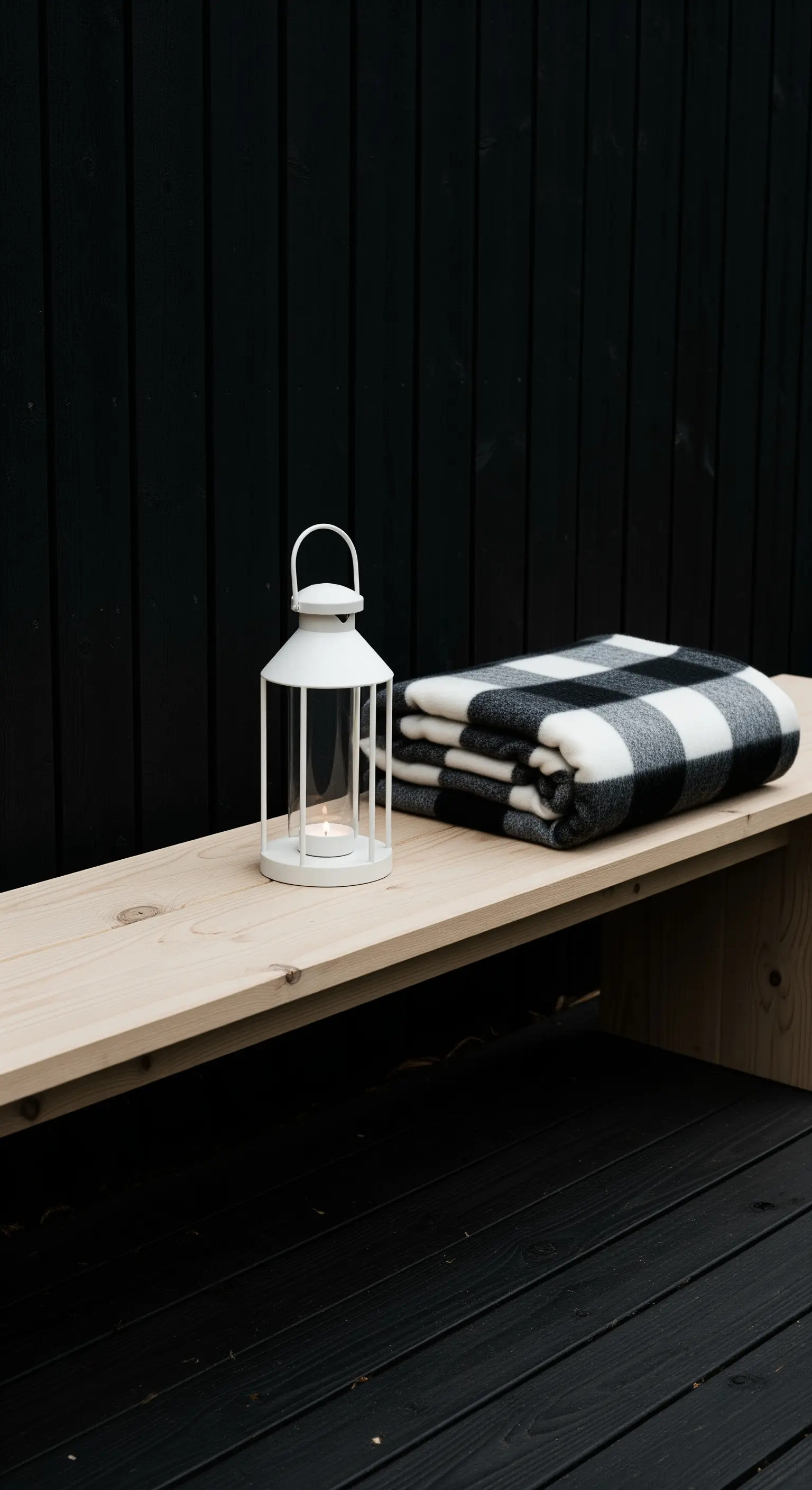 Panca in legno chiaro con lanterna bianca e plaid, contro una parete di legno nero.