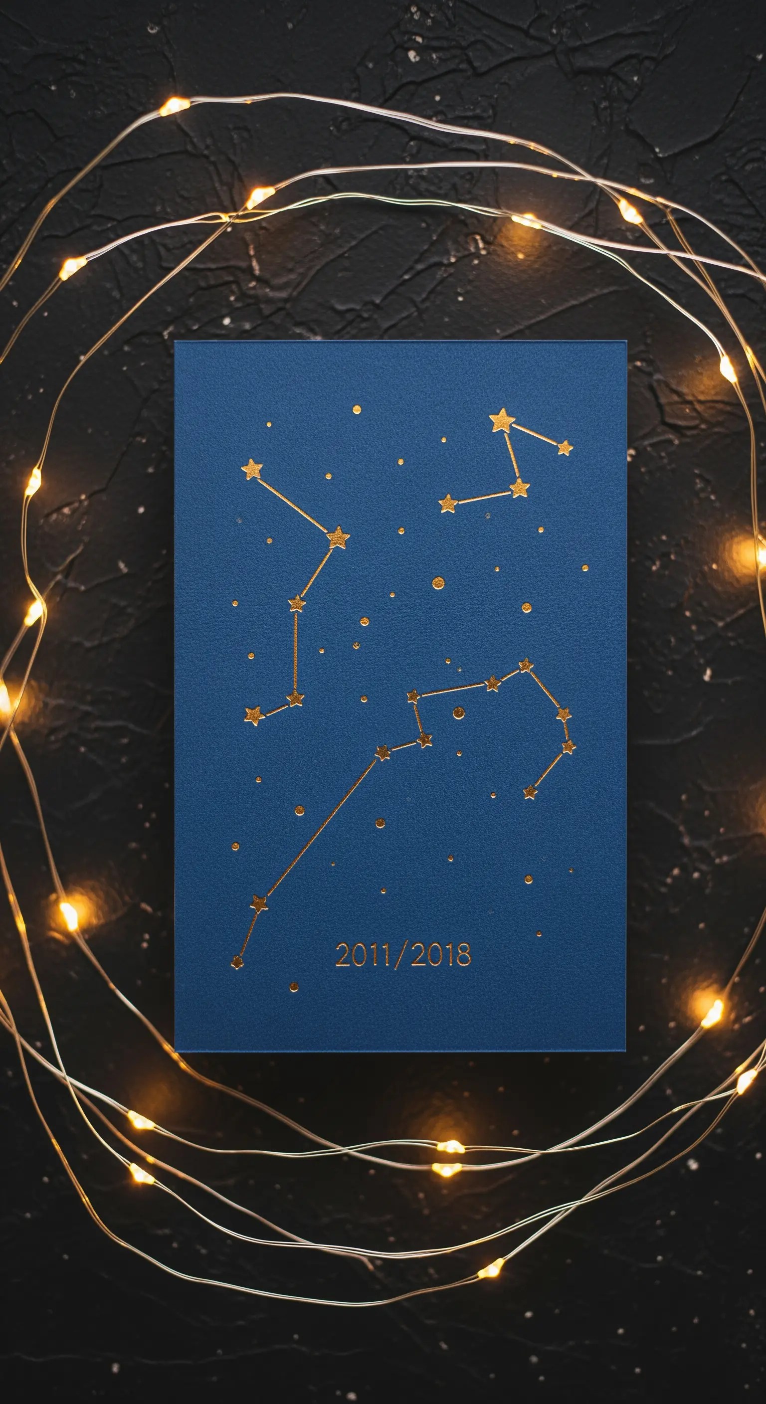 Cartoncino blu con la mappa delle costellazioni dorata e due date, circondato da lucine.