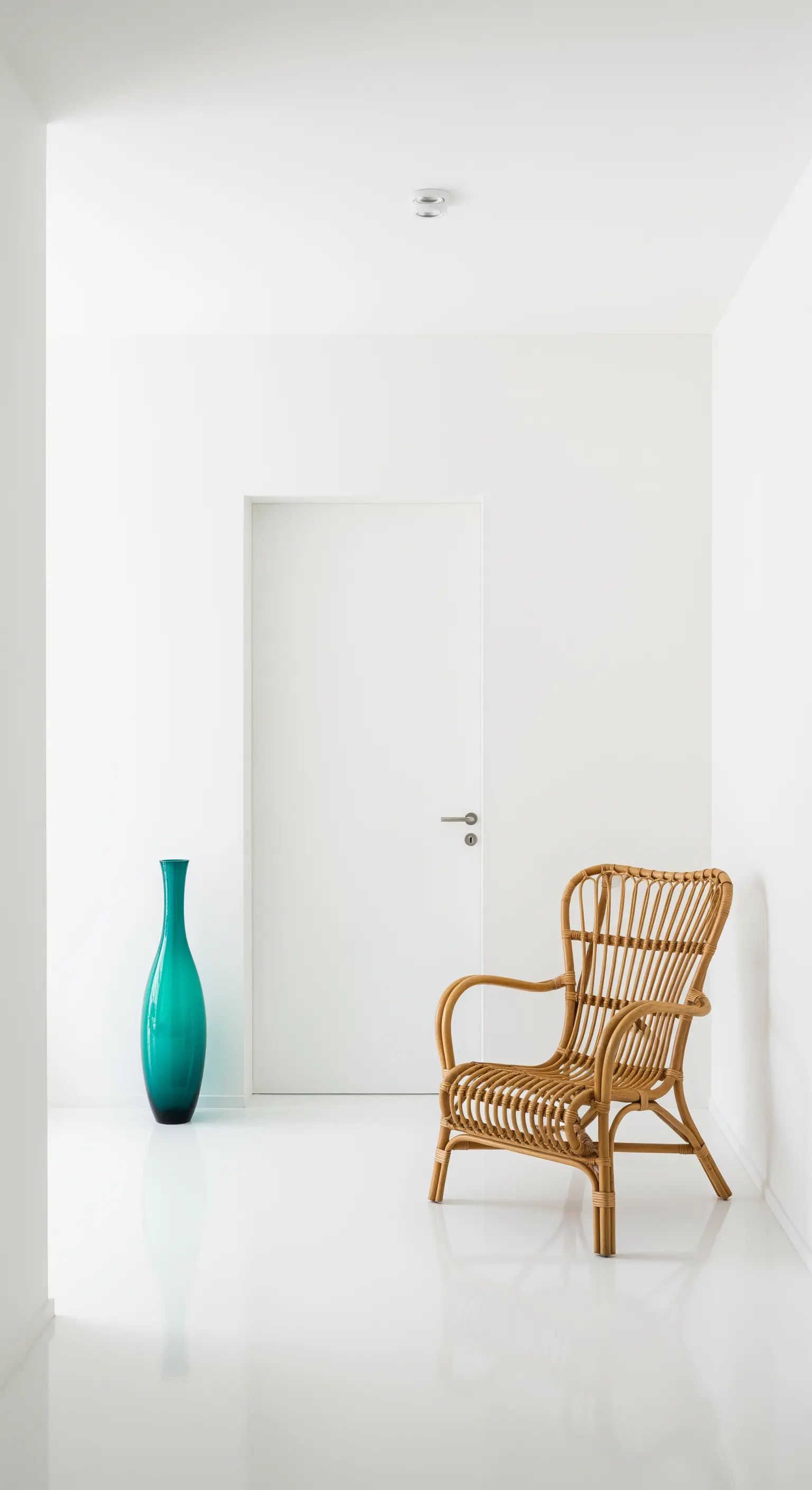 Corridoio bianco minimalista con una sedia in rattan e un vaso di vetro verde acqua.