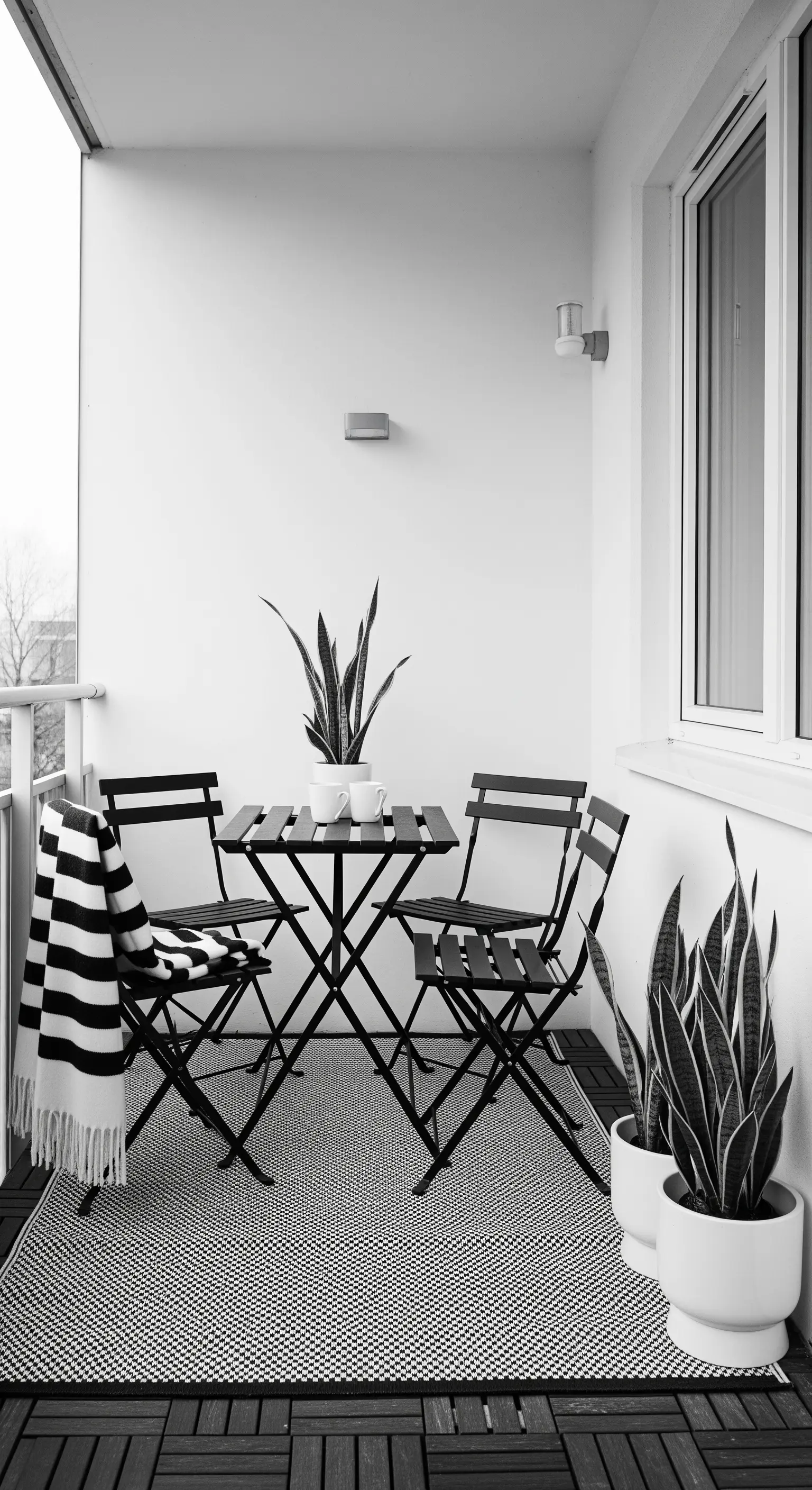 Balcone minimalista in bianco e nero con set da bistrò, piante e coperta a righe.