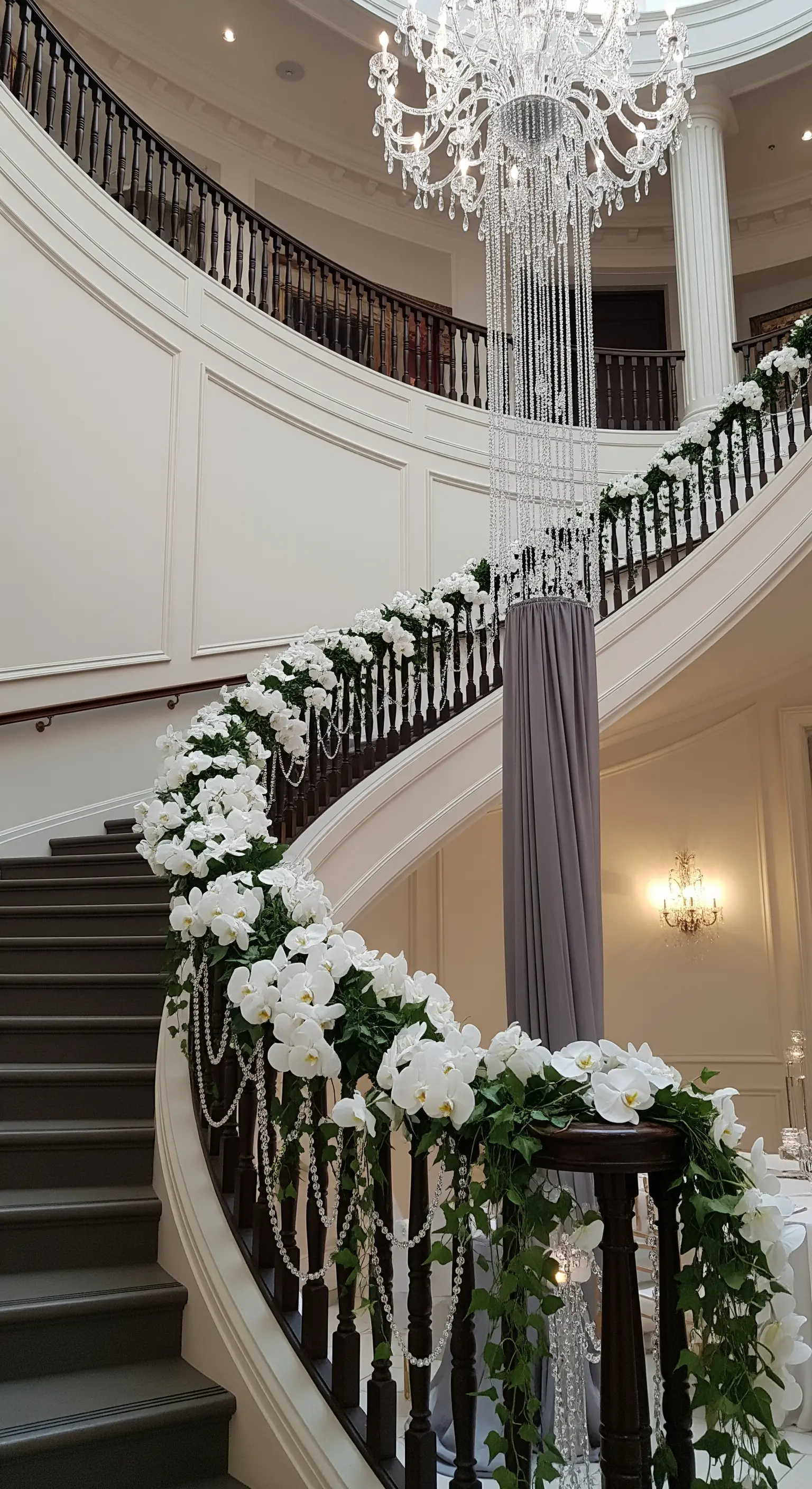 Scalinata curva decorata con una ghirlanda di orchidee bianche e cristalli, con un chandelier sospeso.