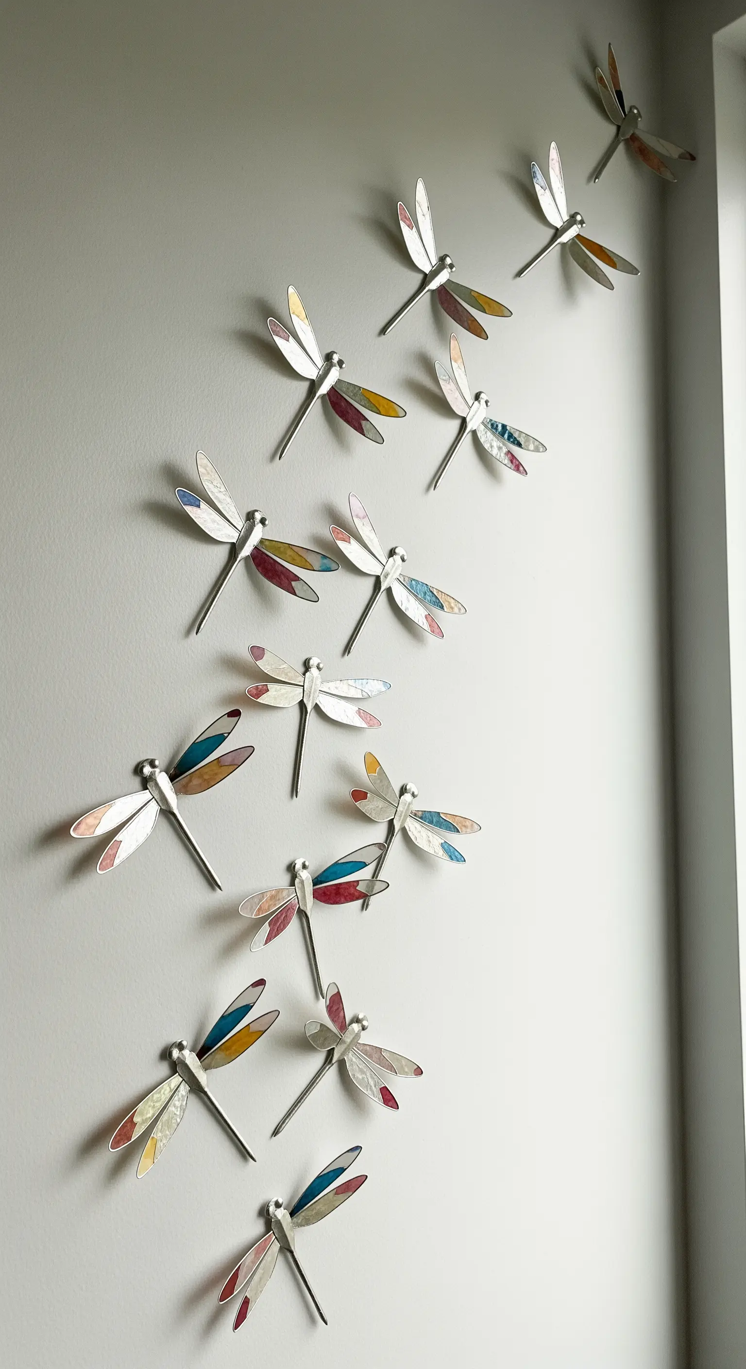 Uno sciame di libellule decorative con ali colorate in stile mosaico, appese a una parete grigia.