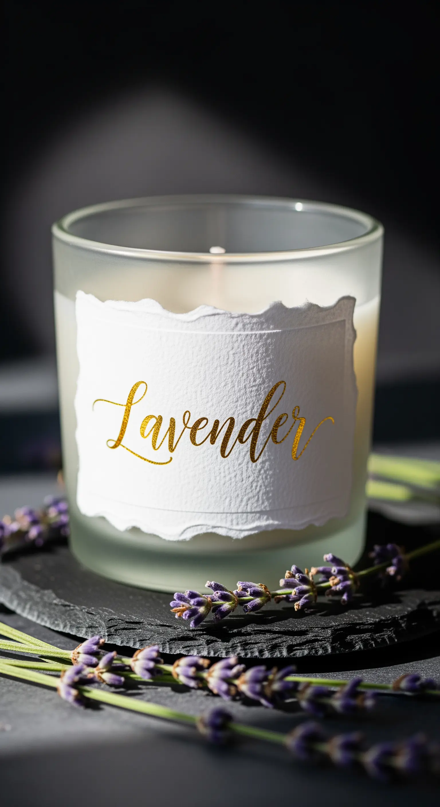 Candela profumata in un vasetto di vetro smerigliato con etichetta dorata 'Lavender', circondata da fiori di lavanda.