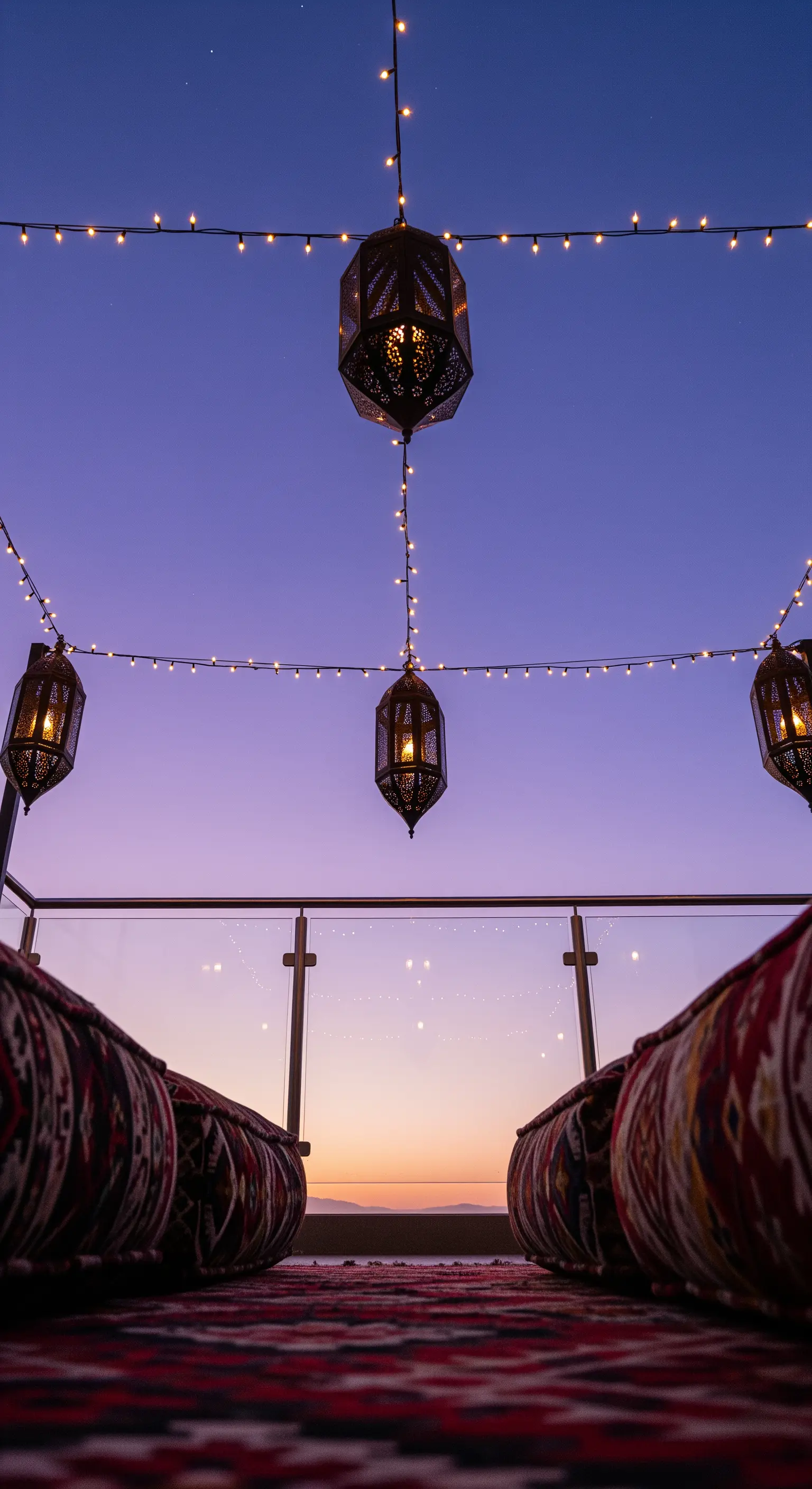 Balcone al tramonto con cuscini bassi su un kilim, lanterne e una fila di lucine.