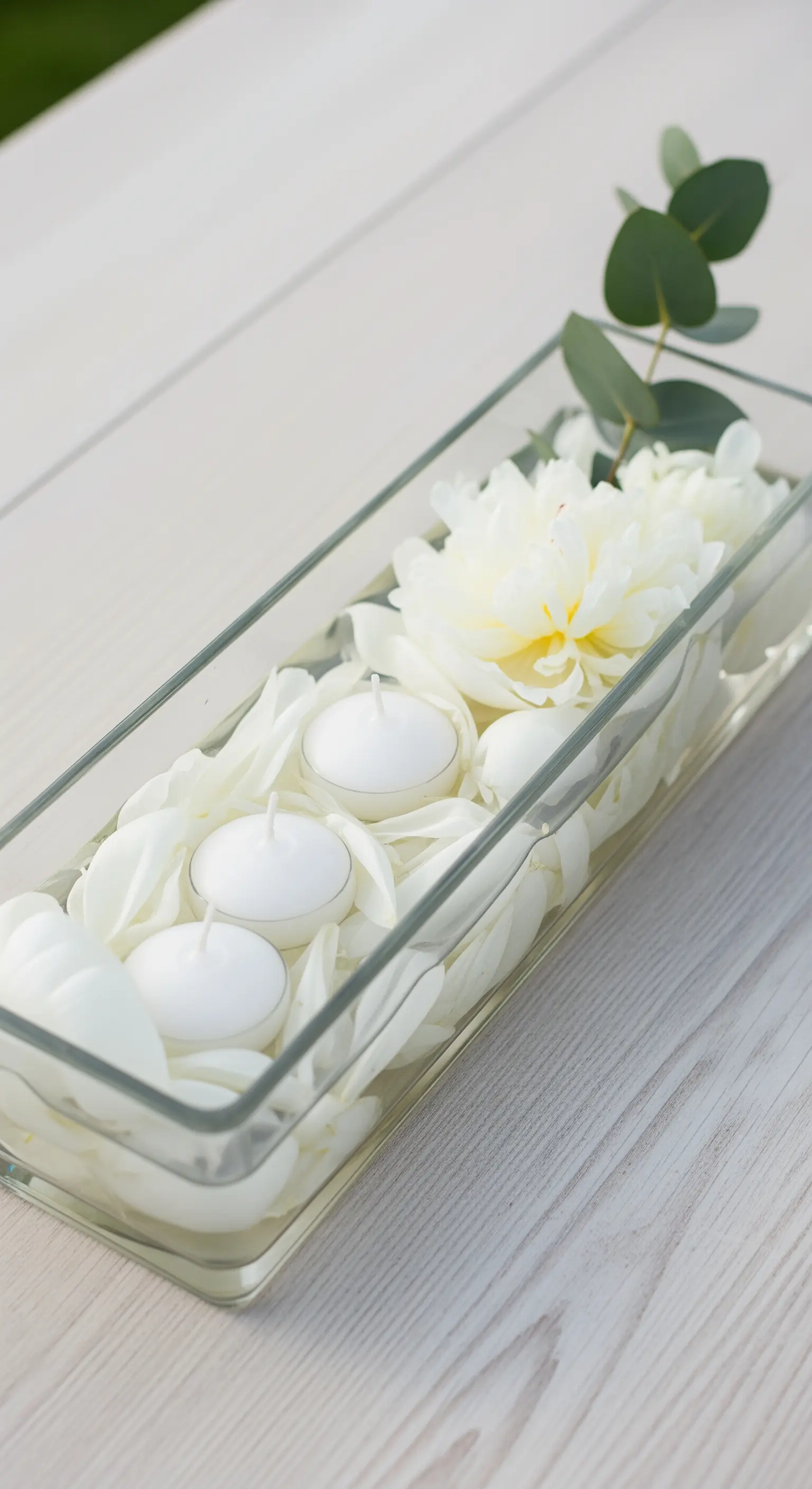 Contenitore in vetro rettangolare con petali bianchi, candele e un ramo di eucalipto.
