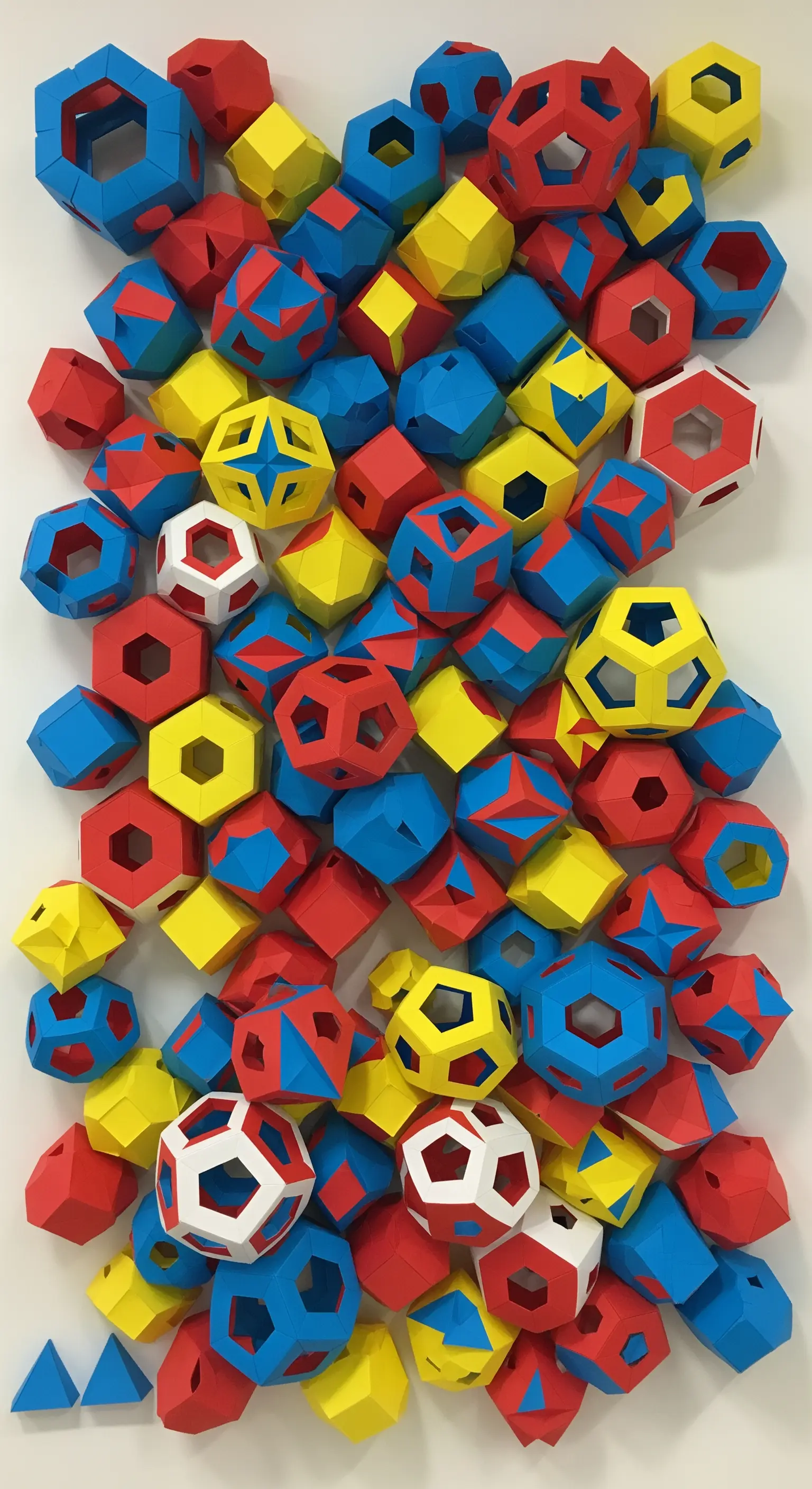 Composizione murale 3D creata assemblando tanti piccoli poliedri di carta di colore rosso, giallo e blu.