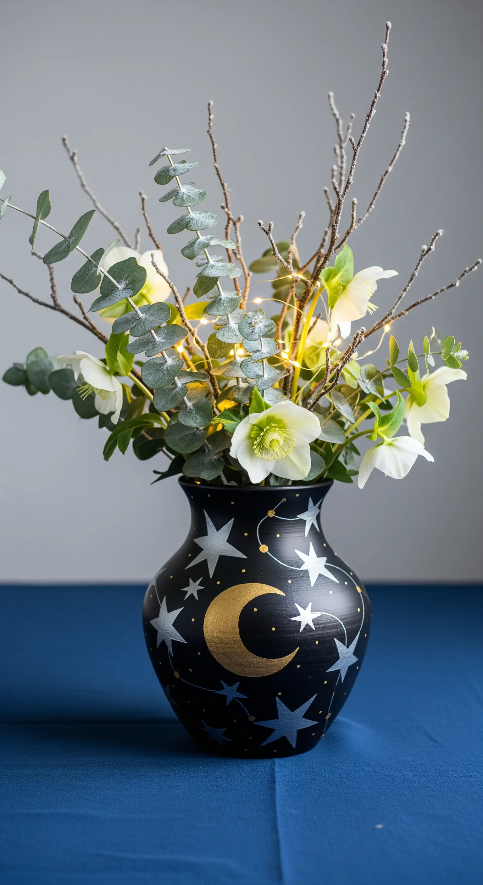 Vaso nero dipinto con luna e stelle dorate, con fiori bianchi e un filo di lucine.
