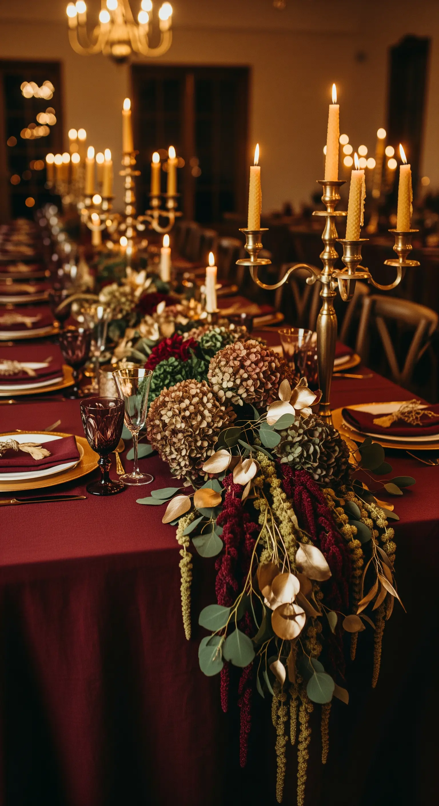 Tavolo formale con tovaglia bordeaux, candelabri dorati e fiori secchi.