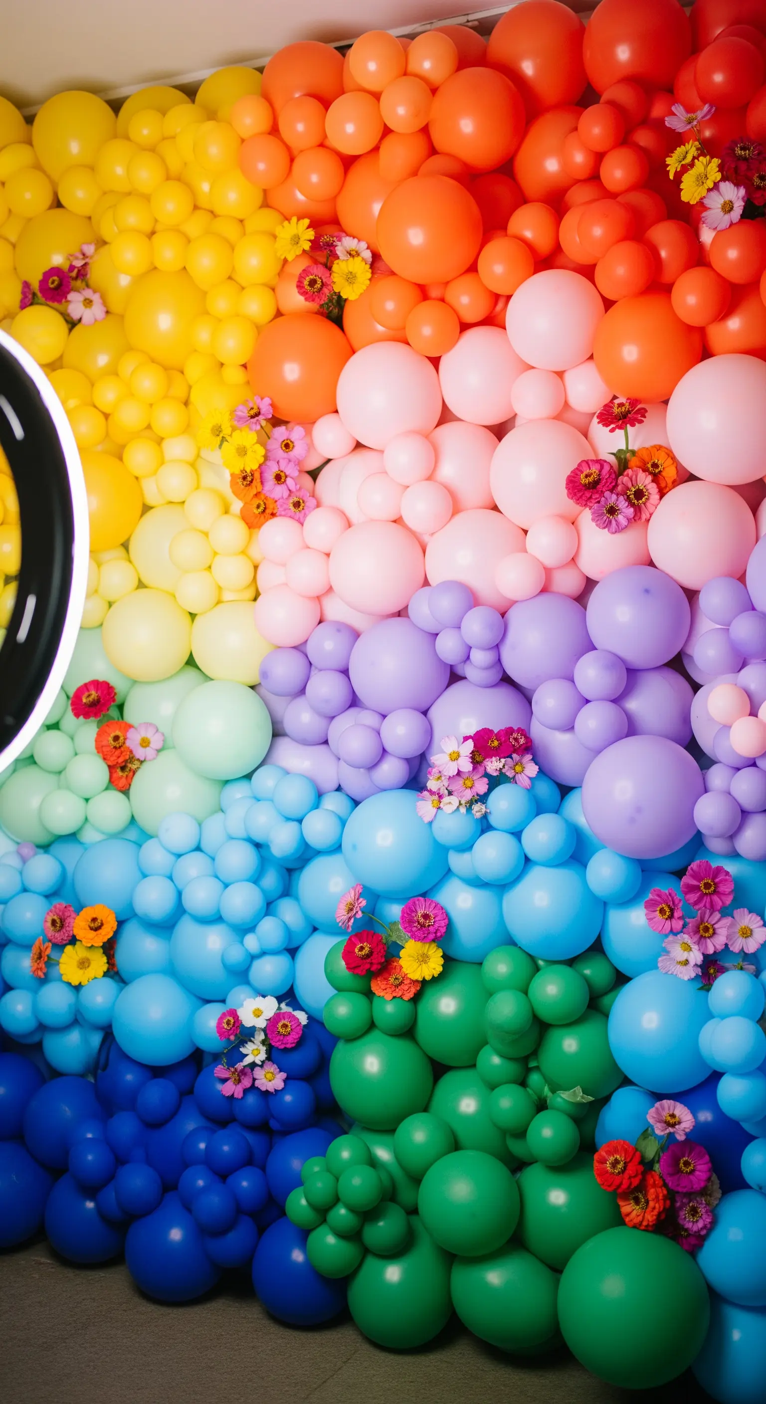 Dettaglio di una parete interamente ricoperta di palloncini arcobaleno con fiori freschi.