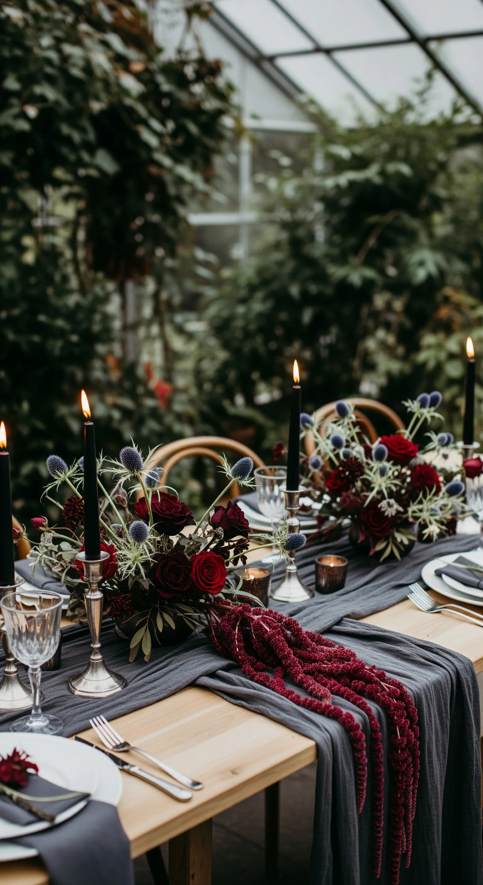 Tavolo in legno con runner grigio scuro, fiori rossi e candele nere in una serra.