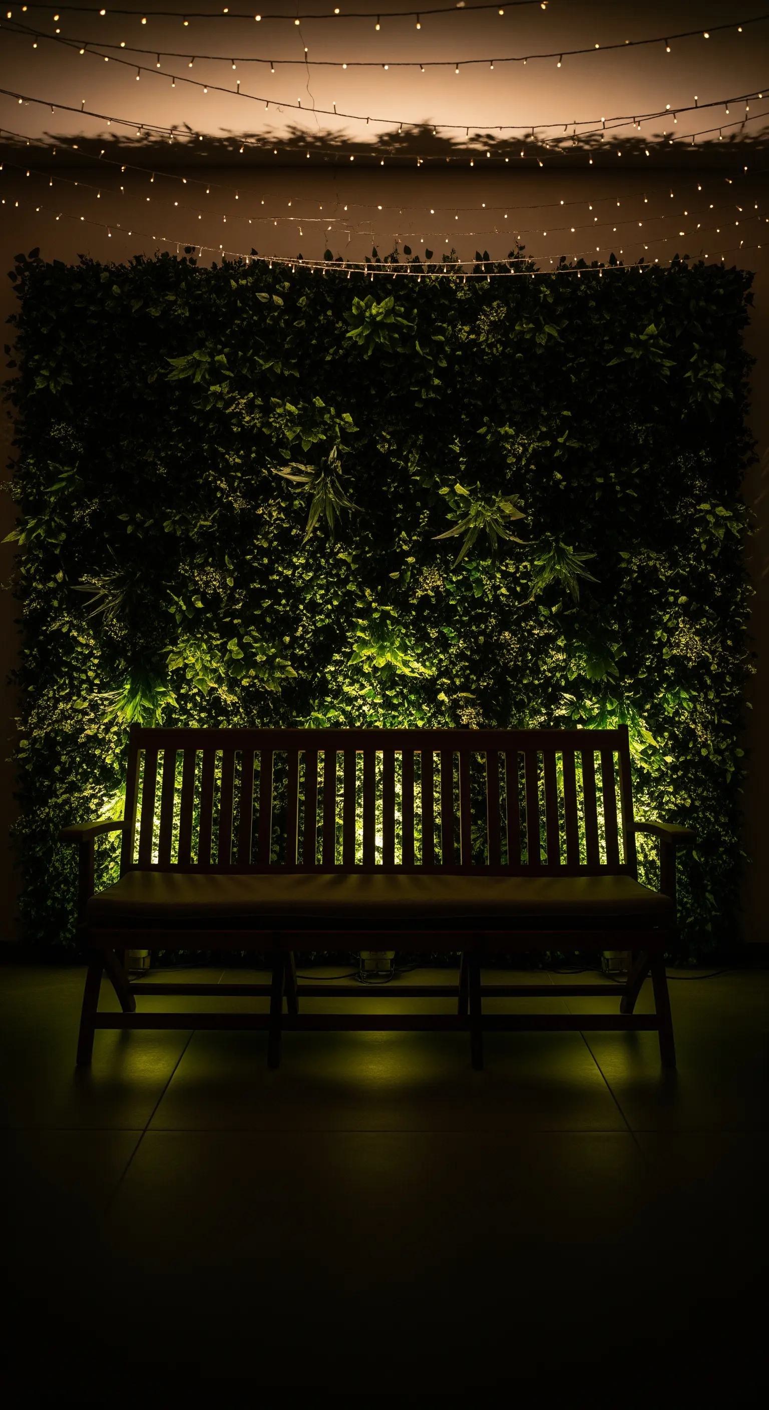 Parete verde illuminata di notte, con una panca in legno e luci a cascata dall'alto.