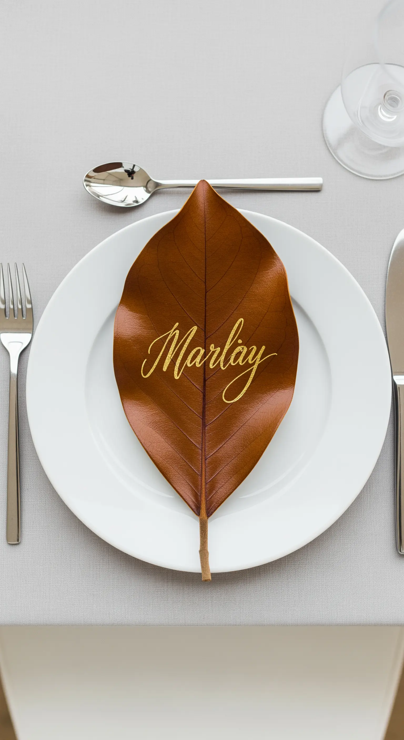 Piatto bianco con una foglia di magnolia secca usata come segnaposto con nome scritto in oro.