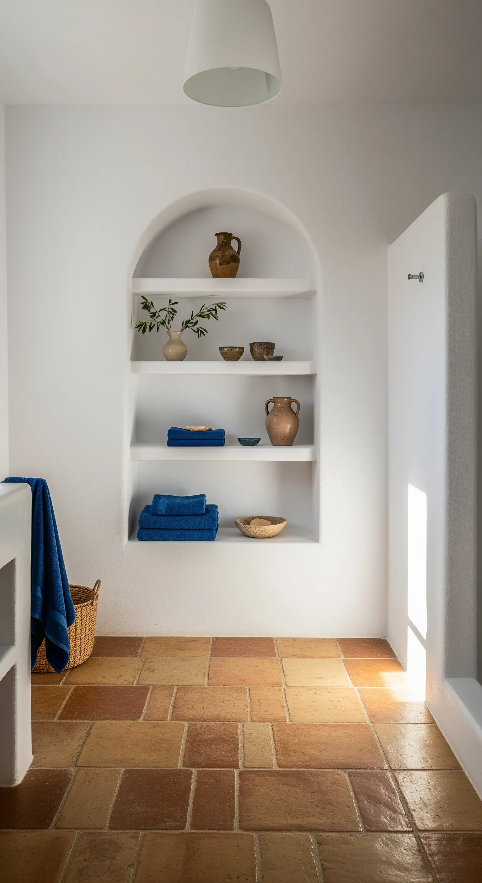 Nicchia bianca ad arco in un bagno con pavimento in cotto, con vasi in ceramica e asciugamani blu.