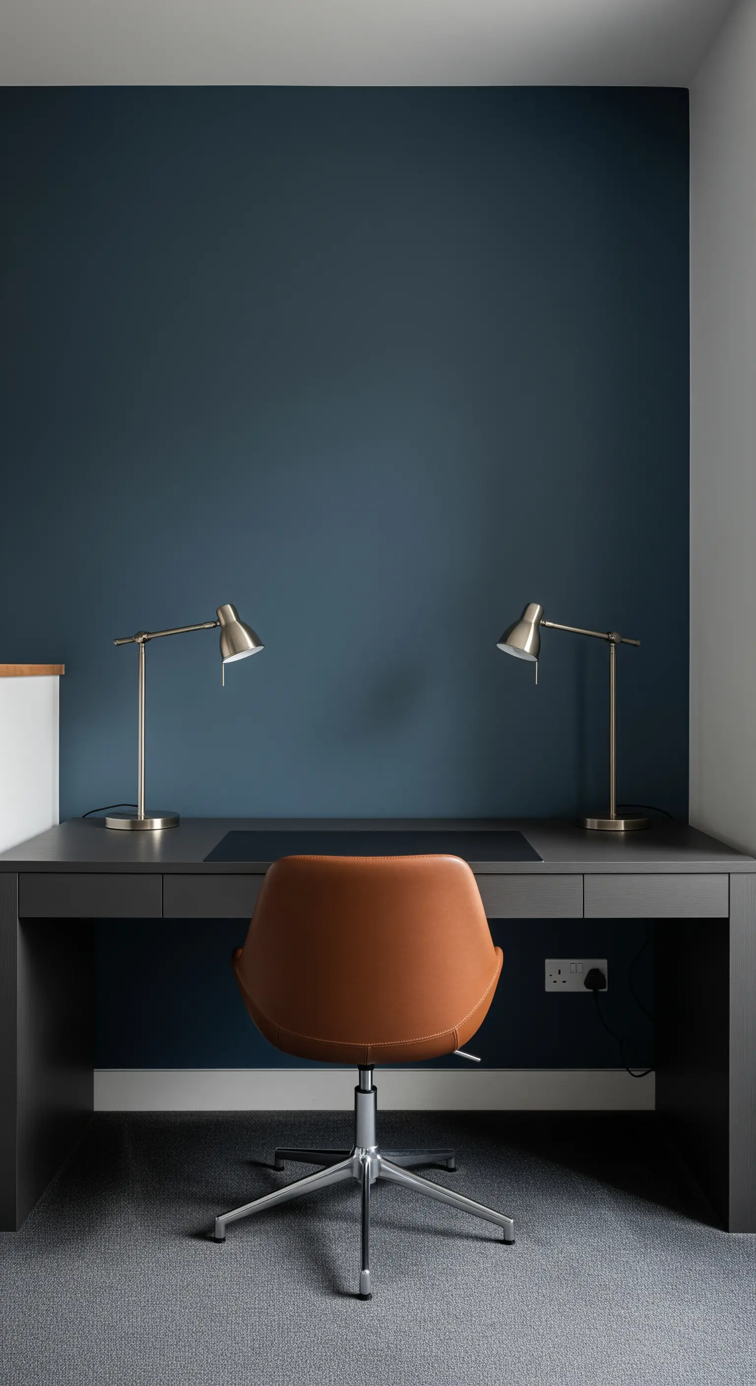 Scrivania grigio scuro contro una parete blu, con sedia in pelle color cuoio e due lampade.