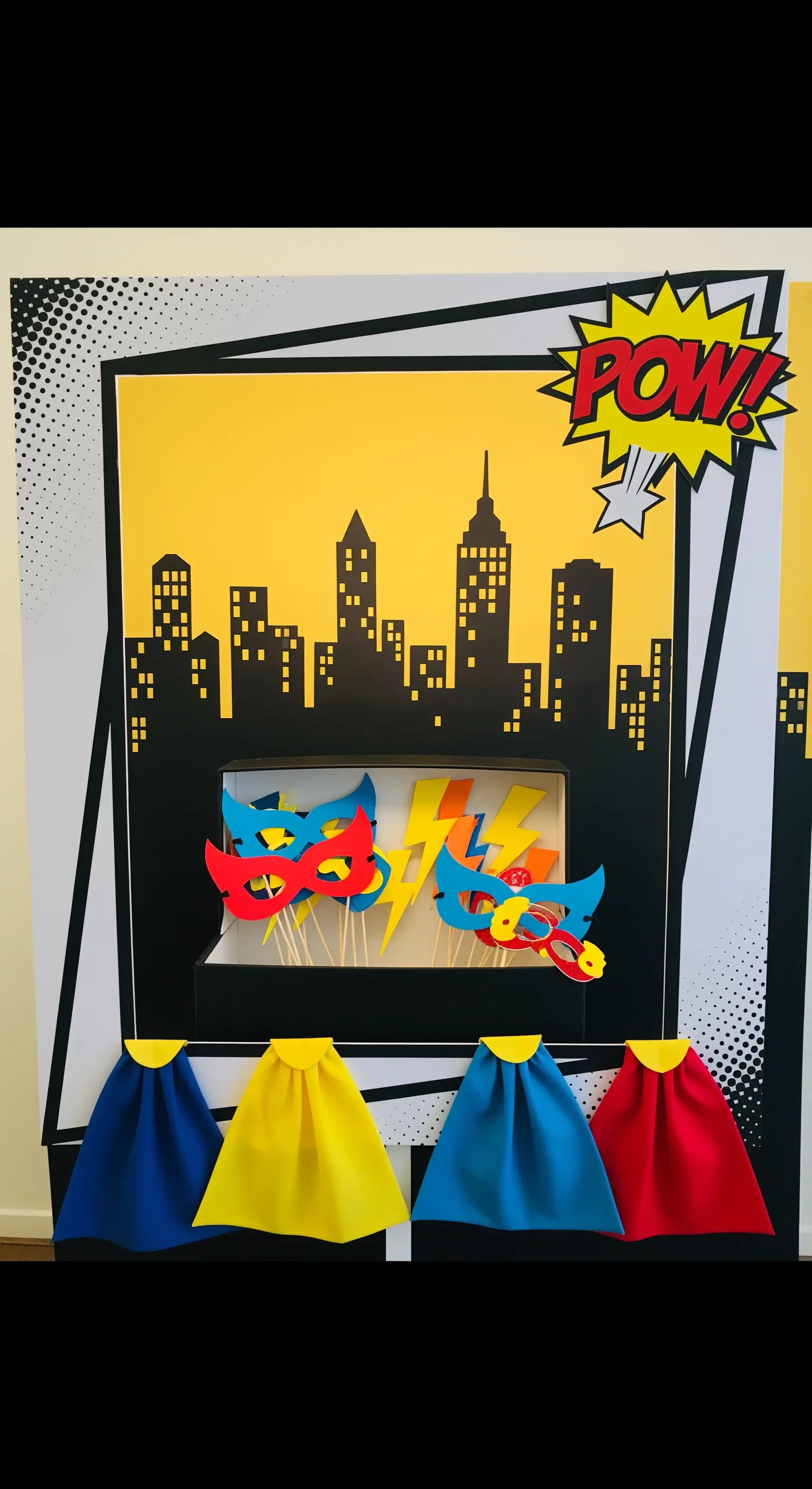 Angolo photobooth a tema supereroi con skyline, maschere colorate e piccoli mantelli.