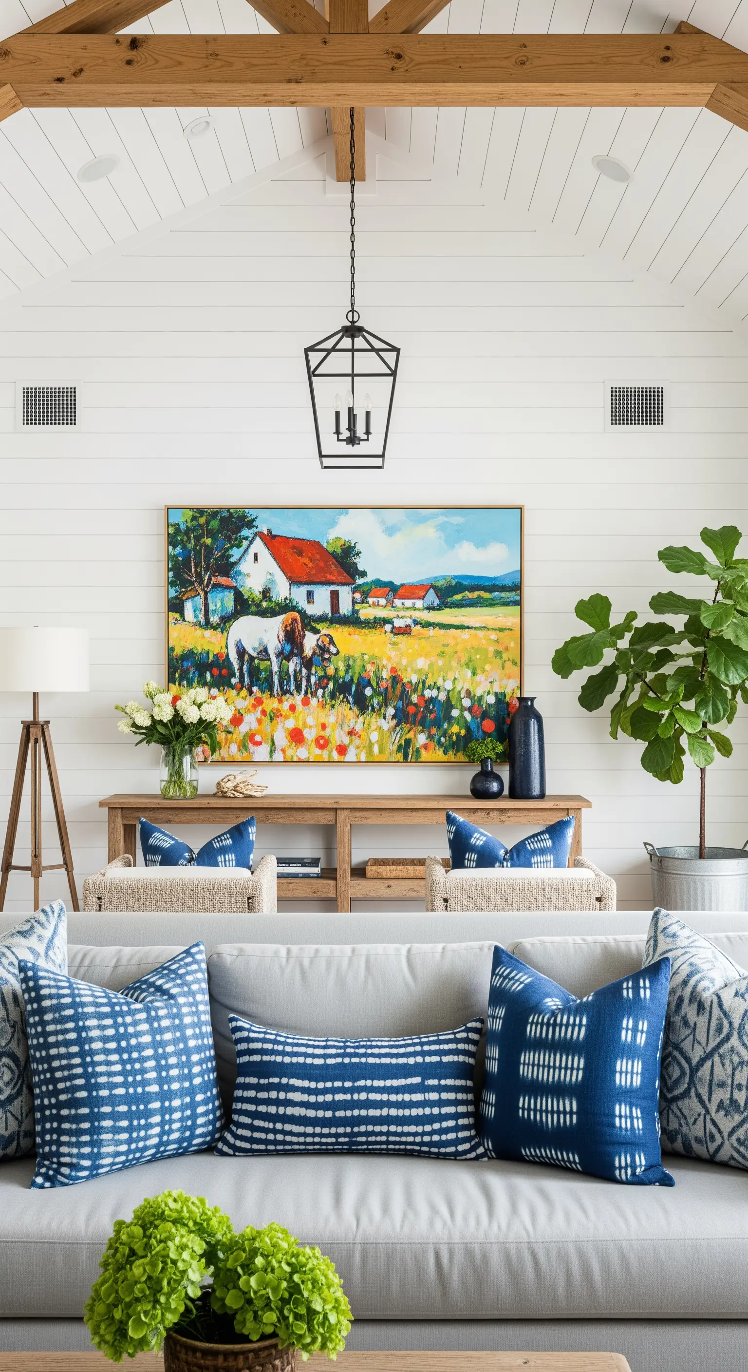 Salotto luminoso in stile farmhouse con pareti bianche, divano grigio con cuscini blu e quadro di campagna.