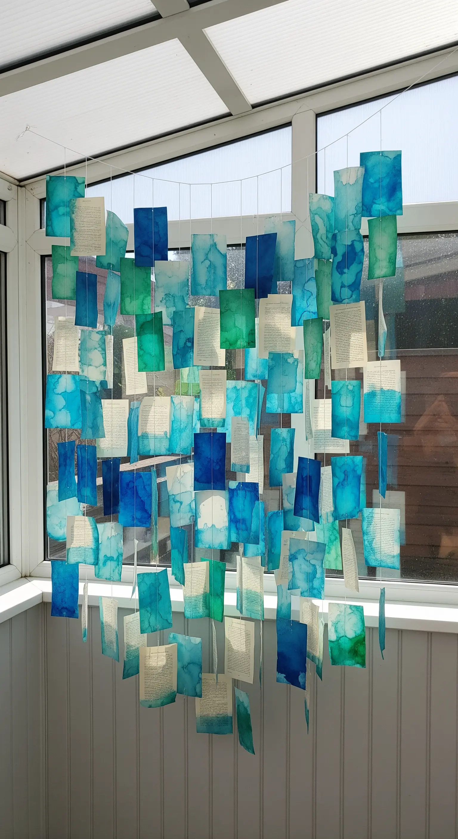Decorazione per finestra creata con pagine di libro dipinte di blu e verde appese a fili.