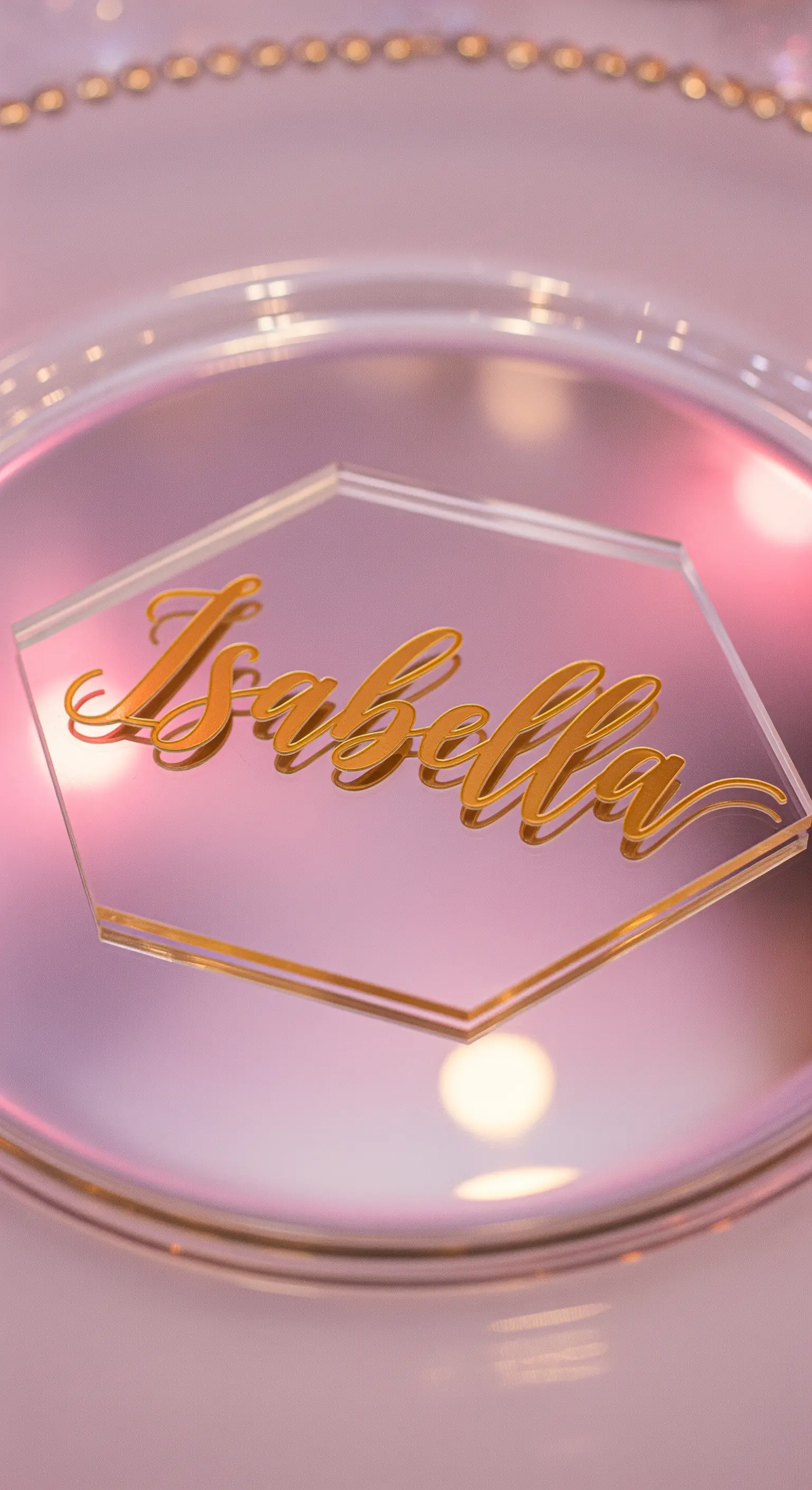 Segnaposto esagonale in acrilico trasparente con nome in rilievo dorato, su un sottopiatto specchiato rosa.