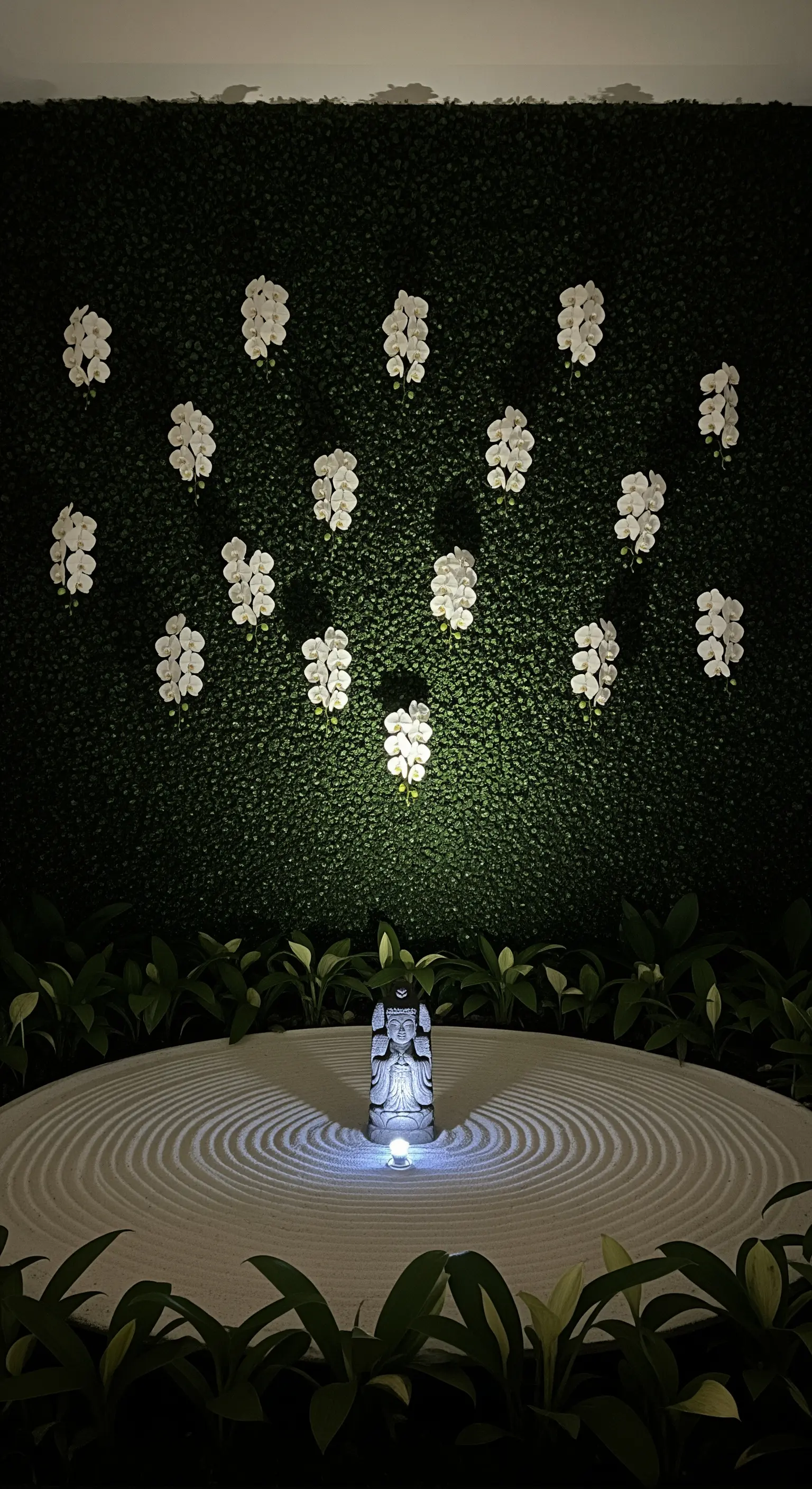 Giardino zen con sabbia bianca, una piccola statua di Buddha e orchidee bianche su una parete verde.