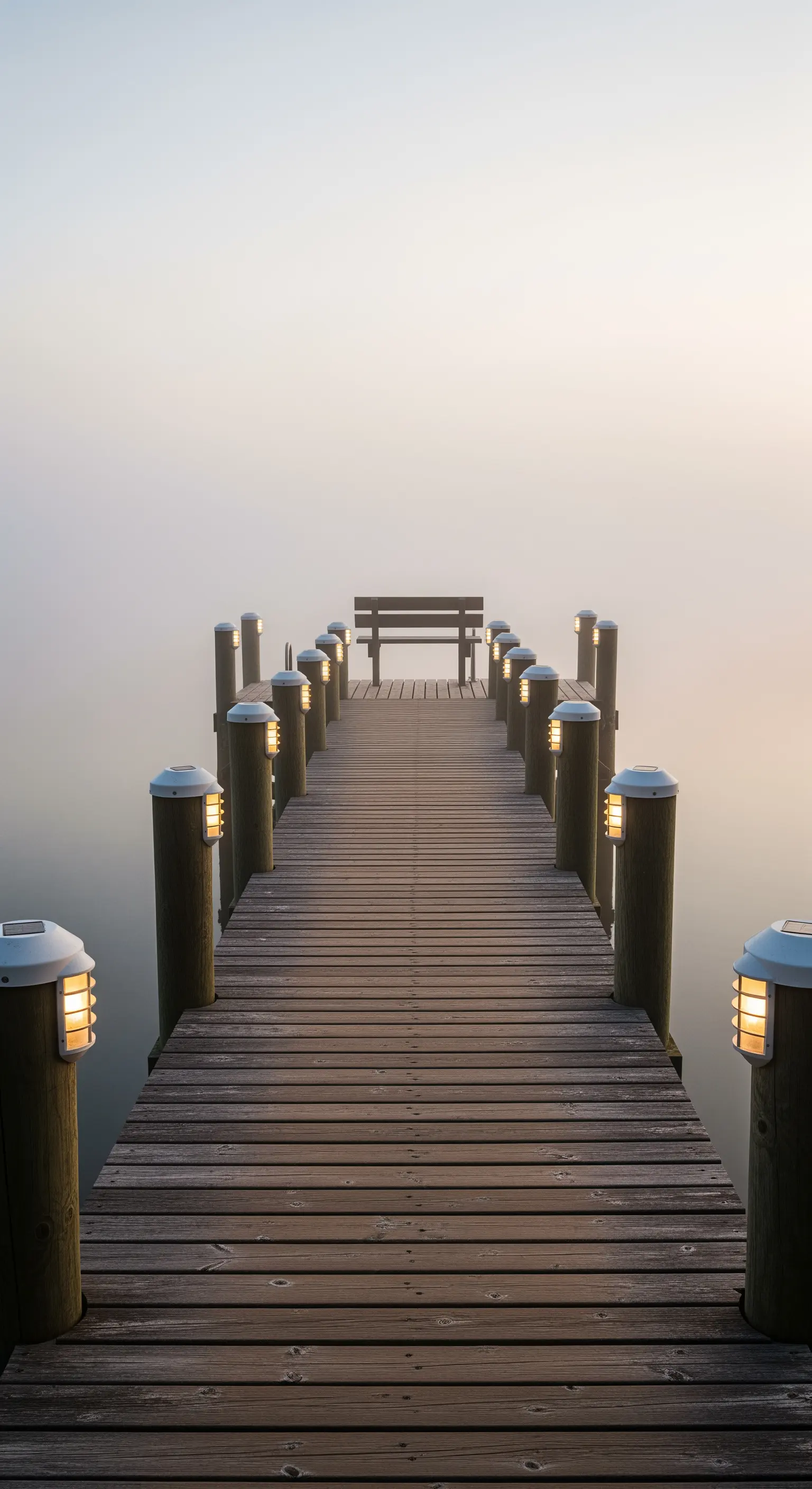Lungo pontile in legno che si estende su un lago nebbioso, illuminato da piccole luci solari sui piloni.