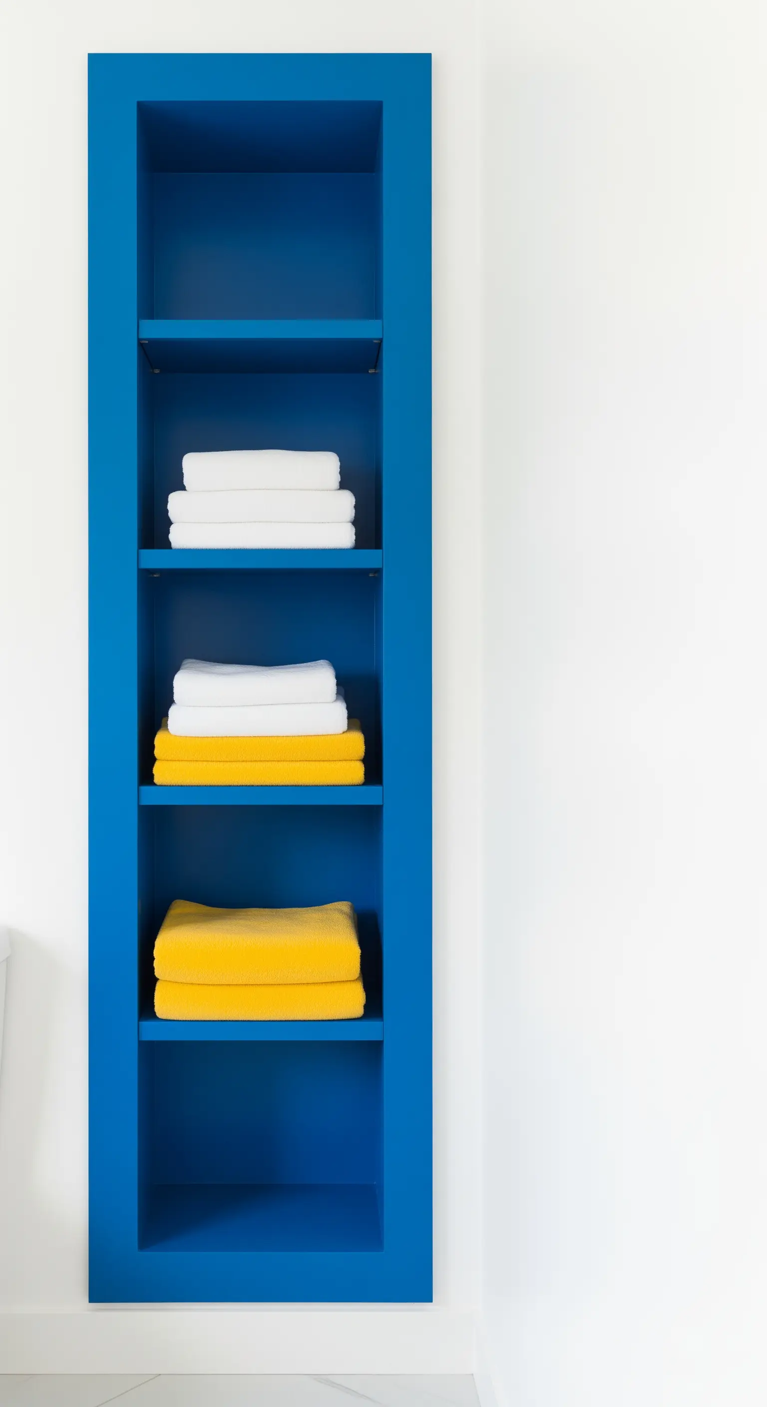 Nicchia a muro dipinta di blu elettrico, con asciugamani bianchi e gialli impilati.