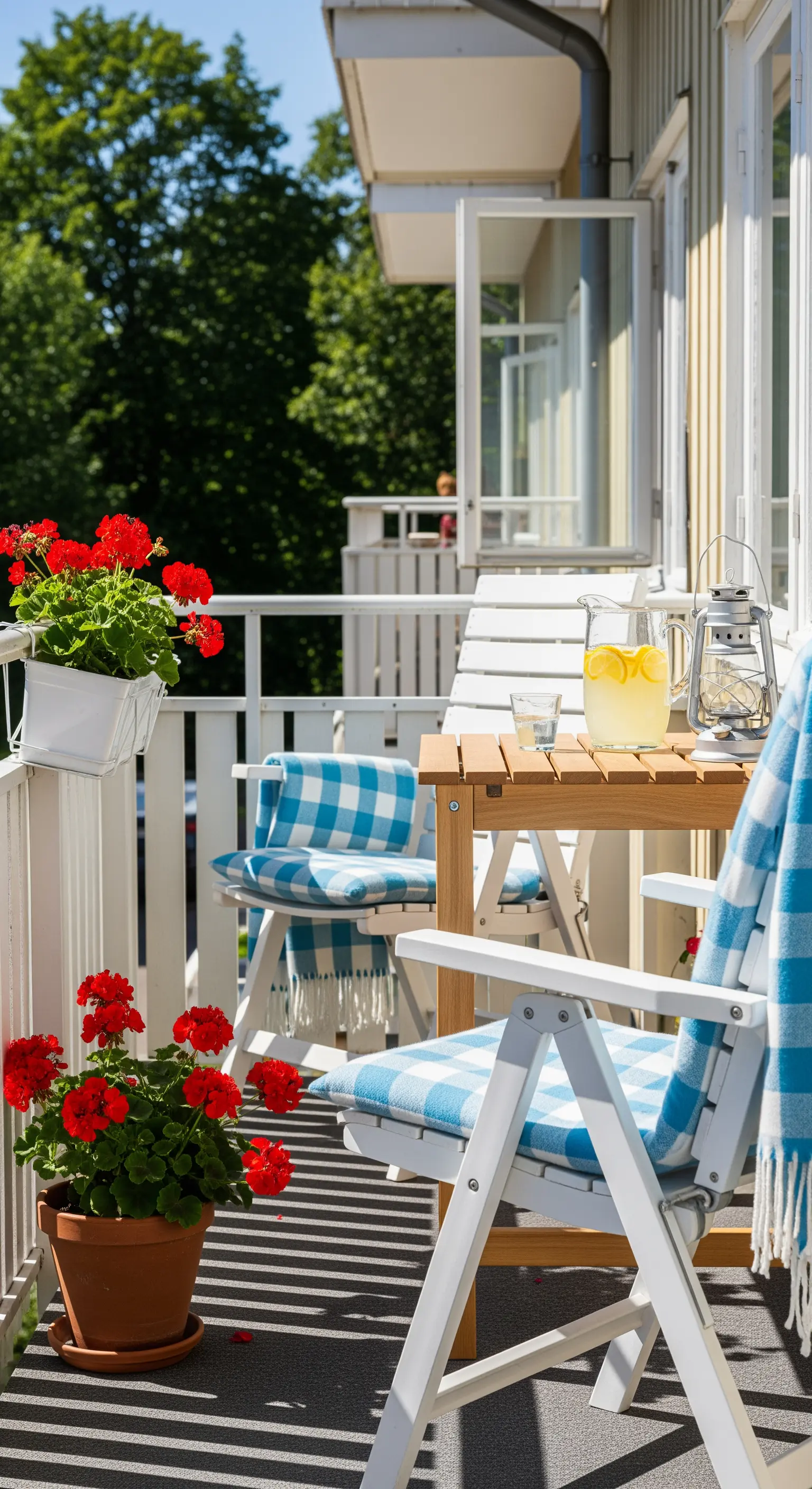 Balcone soleggiato con sedie in legno bianco, cuscini a quadri blu e gerani rossi.