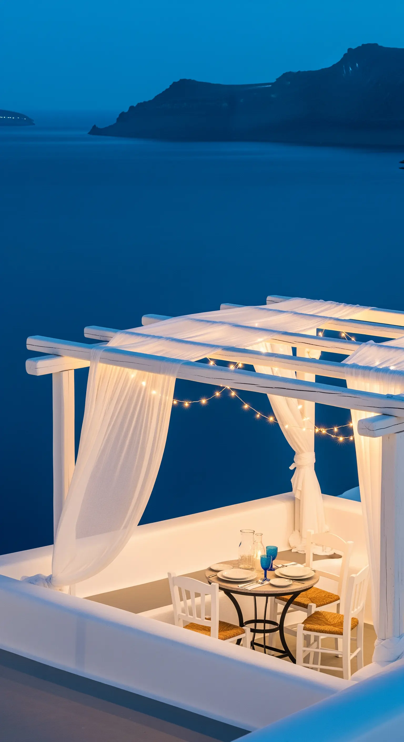 Pergolato bianco con tende leggere e fili di luce affacciato sul mare al crepuscolo.