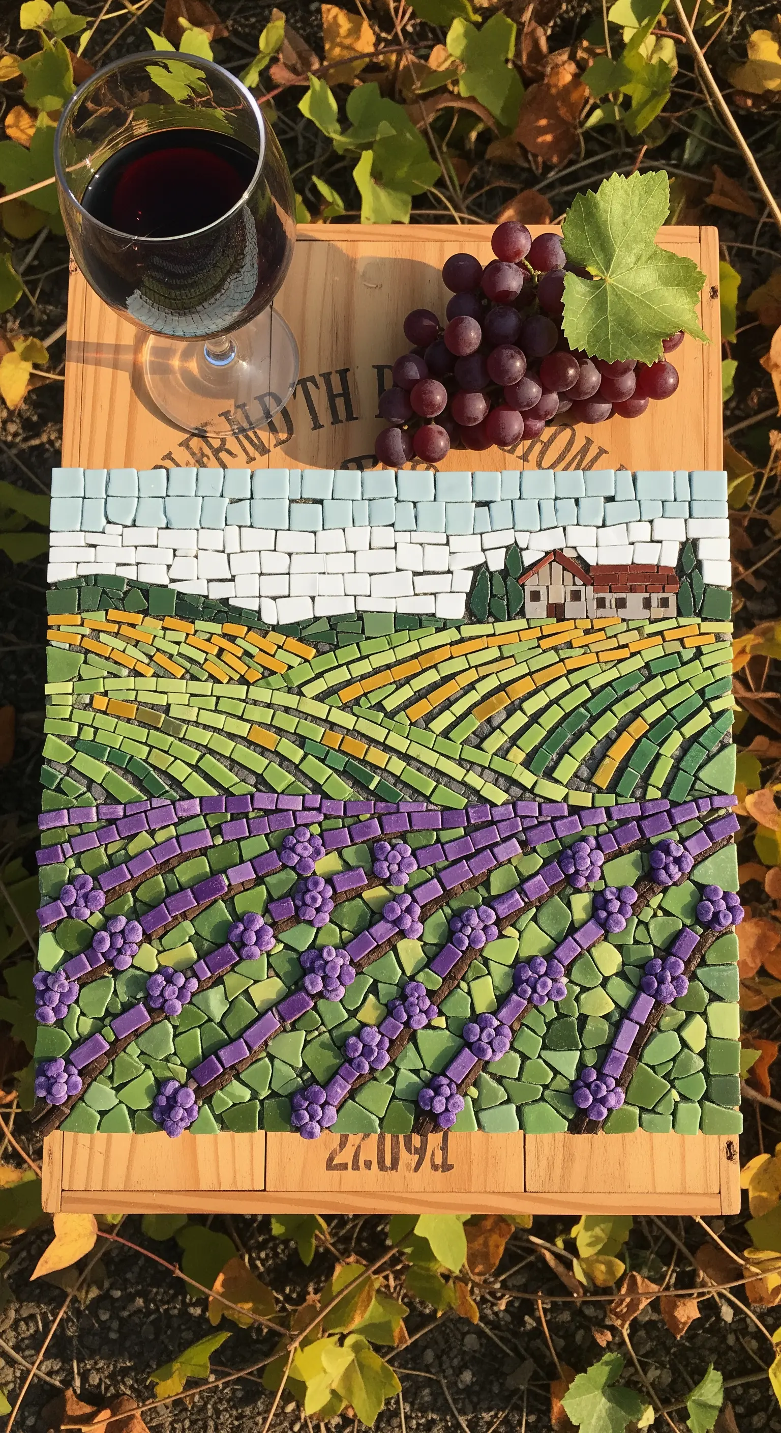 Mosaico di un paesaggio collinare con vigneti viola e verdi, esposto su una cassetta di legno.