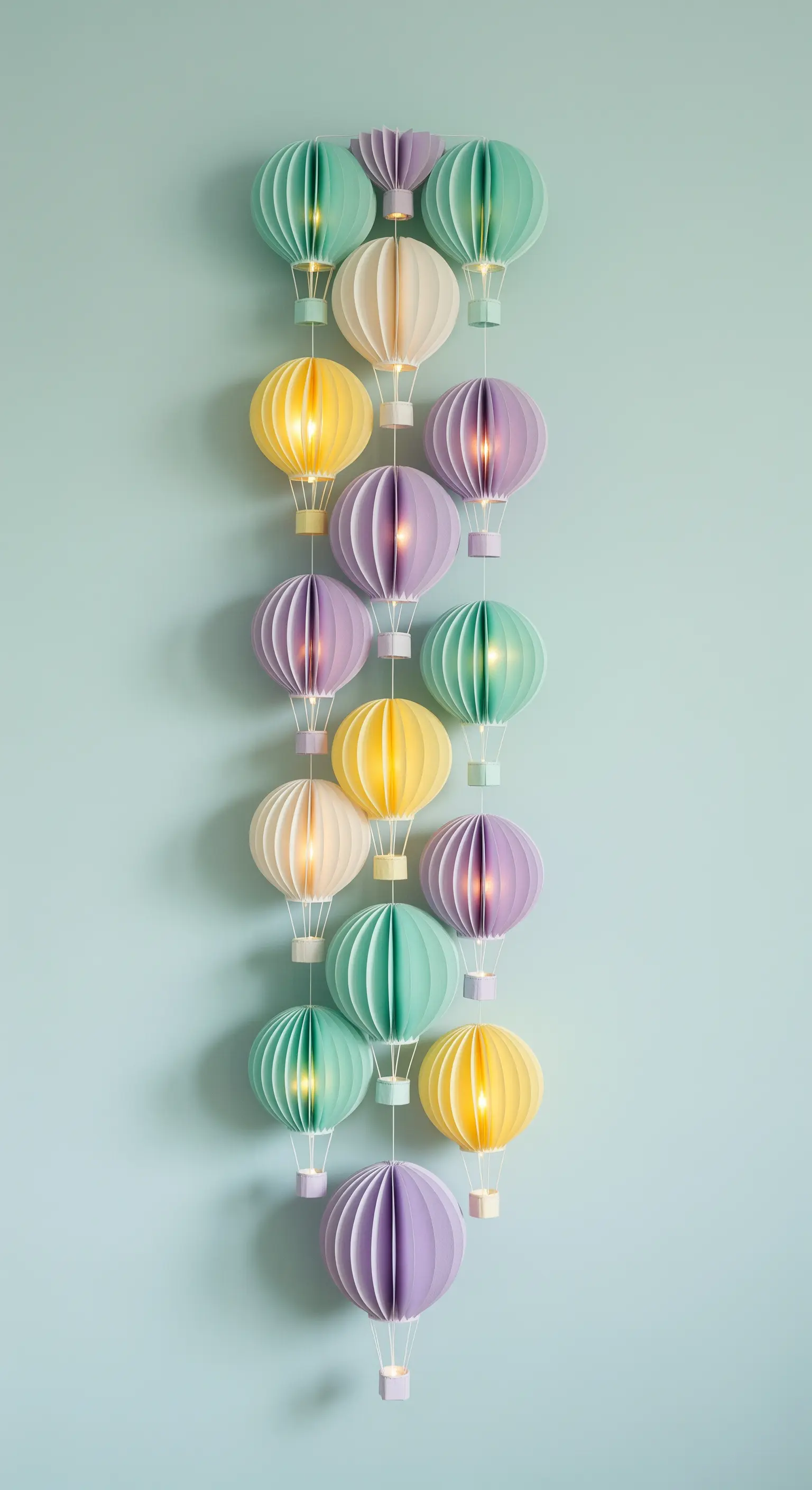 Ghirlanda verticale di piccole mongolfiere di carta illuminate in colori pastello su parete azzurra.